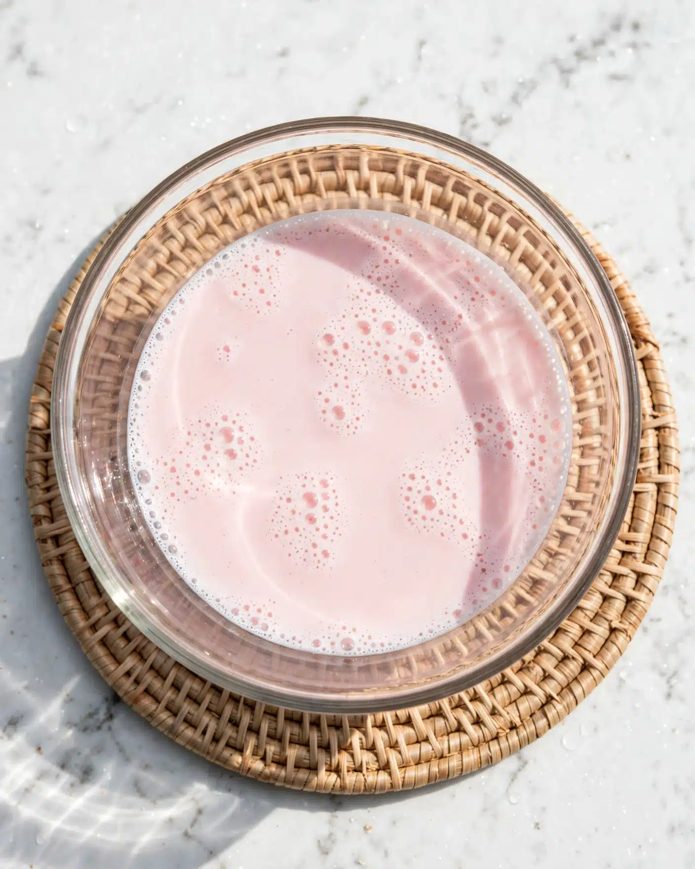 Mohabbat Ka Sharbat | Watermelon Rose Drink Recipe 4 The image shows a clear glass bowl filled with a smooth, pale pink liquid that has small bubbles scattered on the surface. The bowl rests on a round woven mat placed on a white marbled texture surface. The liquid has a creamy look with tiny frothy spots, and the glass bowl allows light to shine through, creating soft reflections. photo taken with an iphone --ar 4:5 --v 7