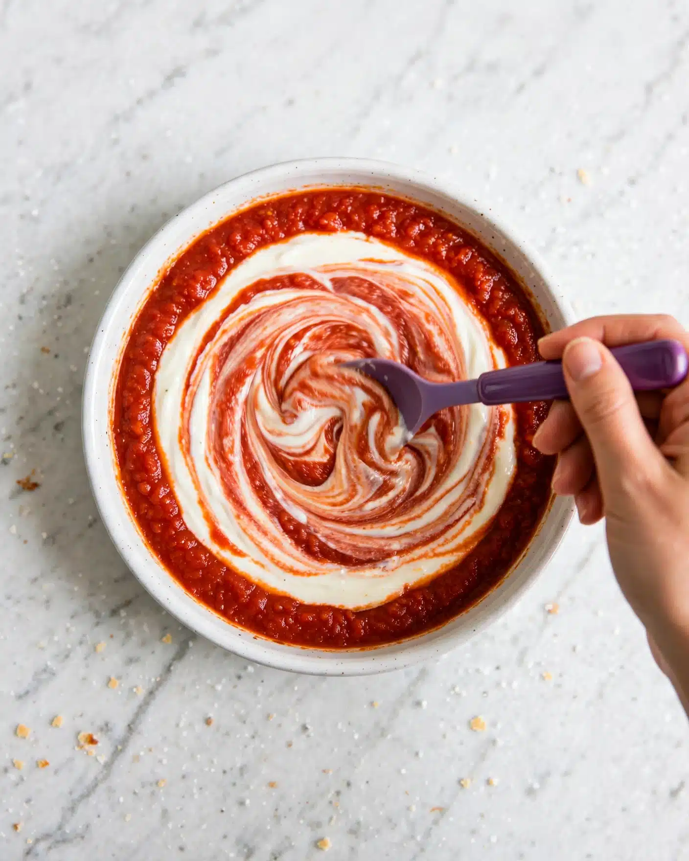 Cottage Cheese Pizza Bowl Recipe 3 A white round dish filled with bright red tomato sauce around the edges, with a creamy white layer swirled in the middle, creating a mixed red and white marbled effect. A woman's hand holds a purple spoon that is swirling the sauce and cream together at the center of the dish. The surface underneath the dish is a white marbled texture. photo taken with an iphone --ar 4:5 --v 7