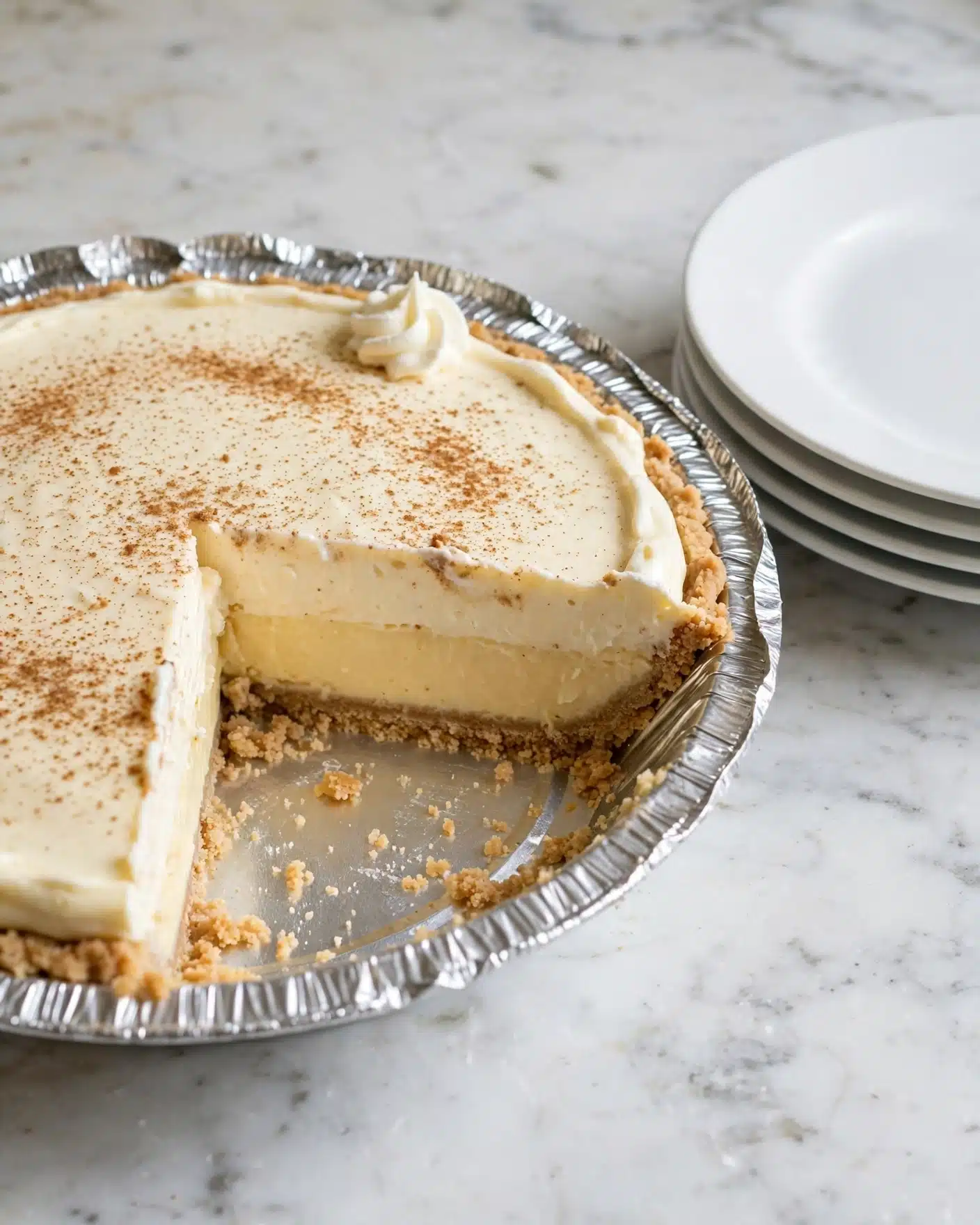 No Bake Eggnog Pie Recipe 5 The image shows a pie in a silver foil pan with one slice missing. The pie has three visible layers: a crumbly light brown crust at the bottom, a thick creamy light yellow middle layer with a smooth texture, and a light creamy top layer sprinkled lightly with brown spice. There is a small swirl of white whipped cream on top near the missing slice. The pie is on a white marbled surface with a stack of white plates nearby. photo taken with an iphone --ar 4:5 --v 7