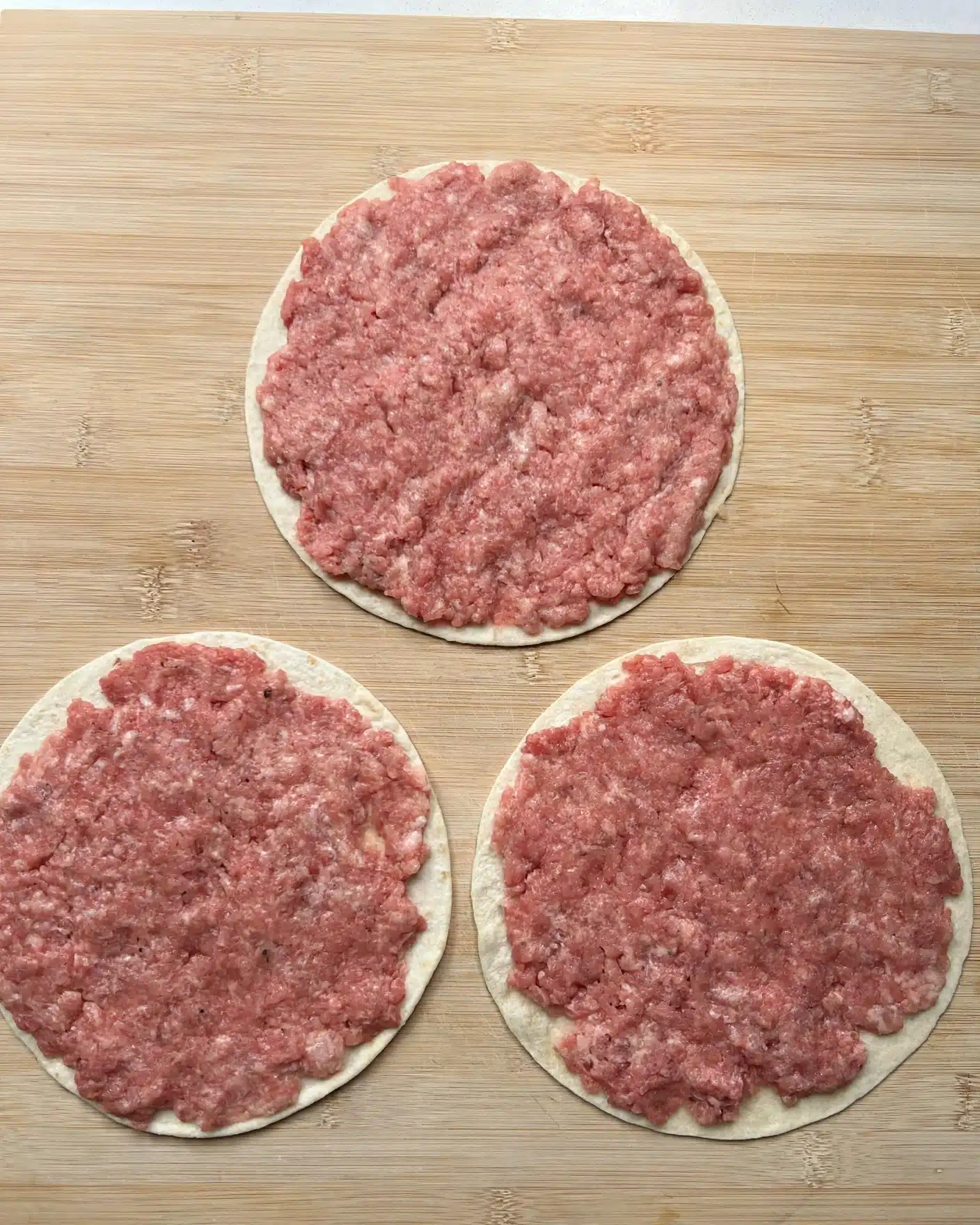 Three round thin dough pieces lay flat on a light wooden board, each topped with a layer of pale pink raw minced meat evenly spread to cover the entire surface. The dough circles have a soft creamy white color and smooth texture visible beneath the meat. The minced meat shows slight ridges and a soft shiny wet look. The arrangement forms a triangle with one circle near the top center and two near the bottom left and right. The natural light highlights the soft textures and subtle color differences between the dough and meat. photo taken with an iphone --ar 4:5 --v 7