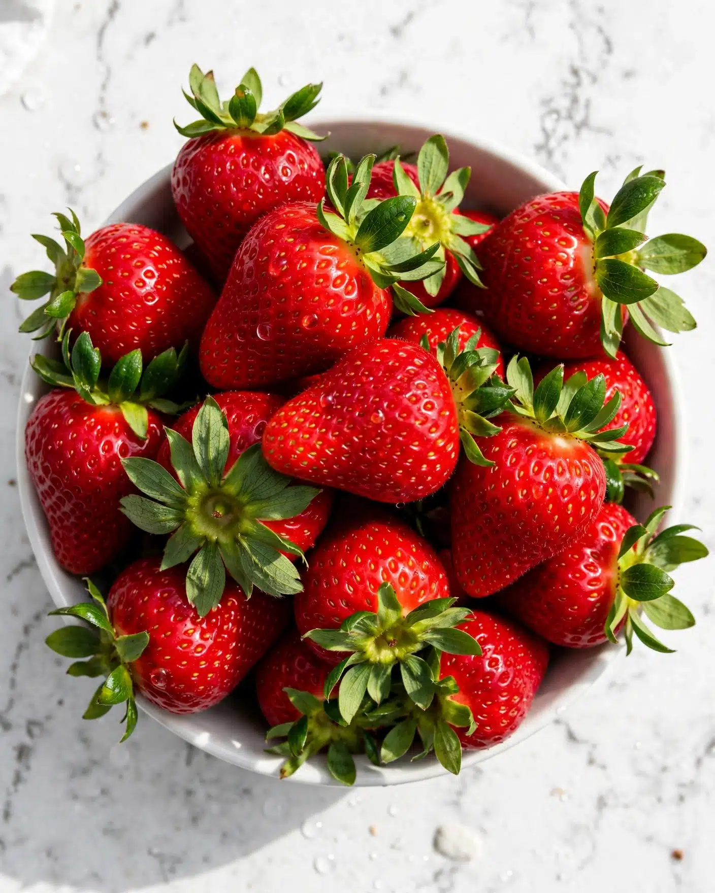 Strawberries and Cream Scones Recipe 4 A close-up view of a bowl filled with bright red strawberries with tiny seeds, each topped with fresh green leaves. The strawberries have a shiny, smooth texture showing their freshness. They fill the white bowl, which sits on a white marbled surface. The image shows a vibrant mix of red and green colors from the fruit and leaves photo taken with an iphone --ar 4:5 --v 7