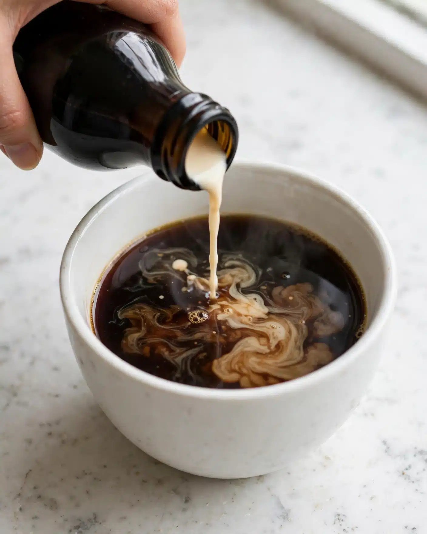 The Best Baileys Chocolate Cookies Recipe 4 A close-up image shows dark black coffee in a white ceramic cup with light cream slowly being poured into it from a dark bottle, creating swirls on the coffee surface. The cup sits on a white marbled surface, and a woman's hand is partially visible holding the bottle at the top left side. The coffee looks smooth and hot, and the cream mixes gently, forming light brown patterns in the dark drink. photo taken with an iphone --ar 4:5 --v 7