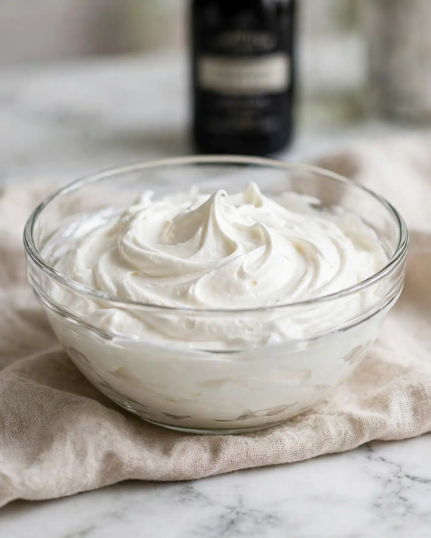 Homemade Cream Horns with Flaky Puff Pastry and Whipped Cream Filling Recipe 4 A clear glass bowl filled with creamy, smooth white whipped mixture that has soft swirls and peaks, showing a light and fluffy texture. The bowl is placed on a light beige textured cloth on a white marbled surface. Behind the bowl, there is a blurred black bottle with a label. The scene is softly lit, highlighting the creamy mixture's texture and slight shine. photo taken with an iphone --ar 4:5 --v 7
