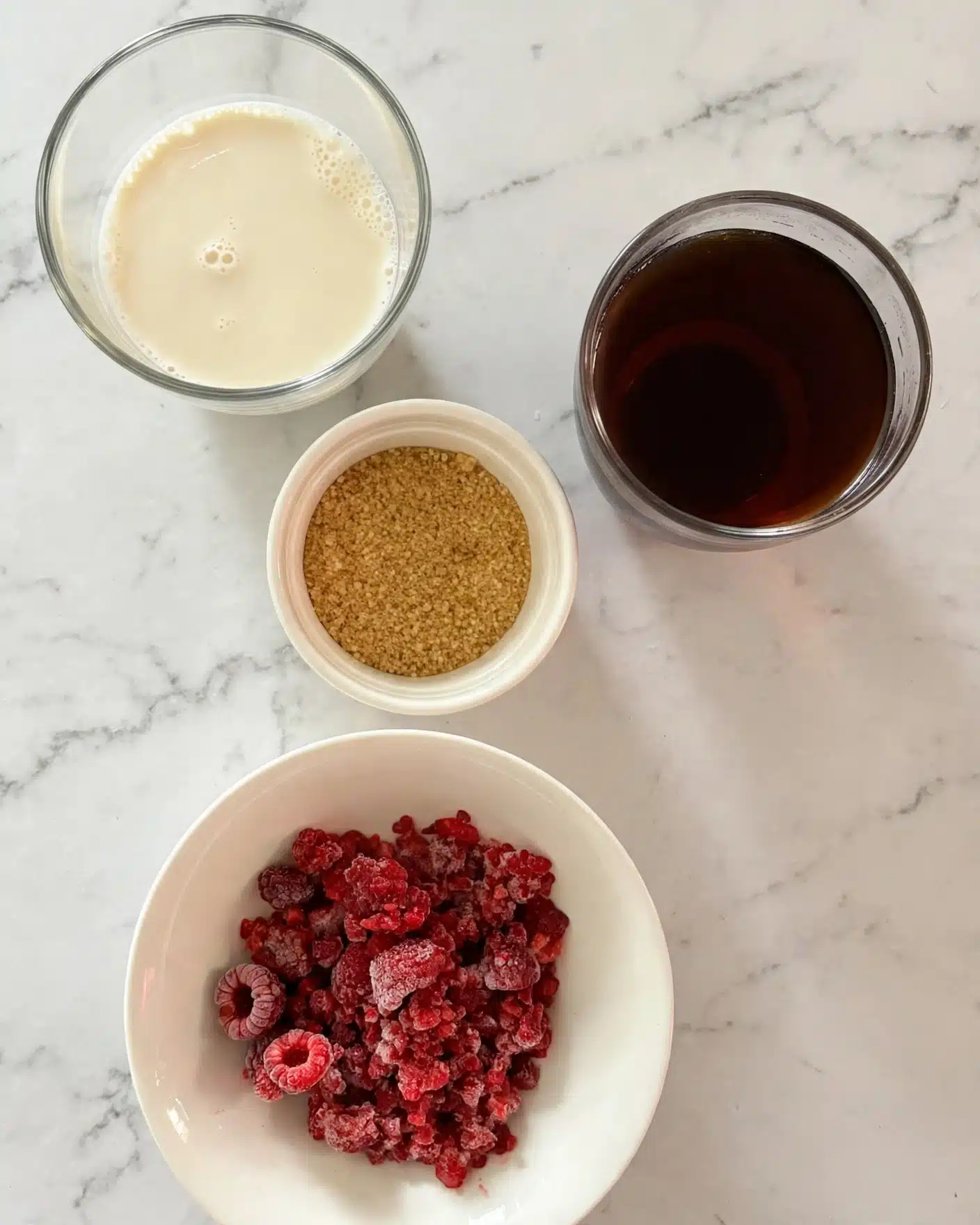The image shows four small bowls placed on a white marbled surface, each containing different ingredients. At the bottom, there is a white bowl filled with bright red frozen raspberries with a frosty texture. Just above it, a small white bowl holds light brown granulated sugar with a rough texture. To the right and slightly above the sugar bowl, there is a small round glass container filled with a dark brown liquid that looks smooth and shiny. To the left of the glass, another clear bowl contains a creamy, off-white liquid that looks smooth with small bubbles on the surface. photo taken with an iphone --ar 4:5 --v 7