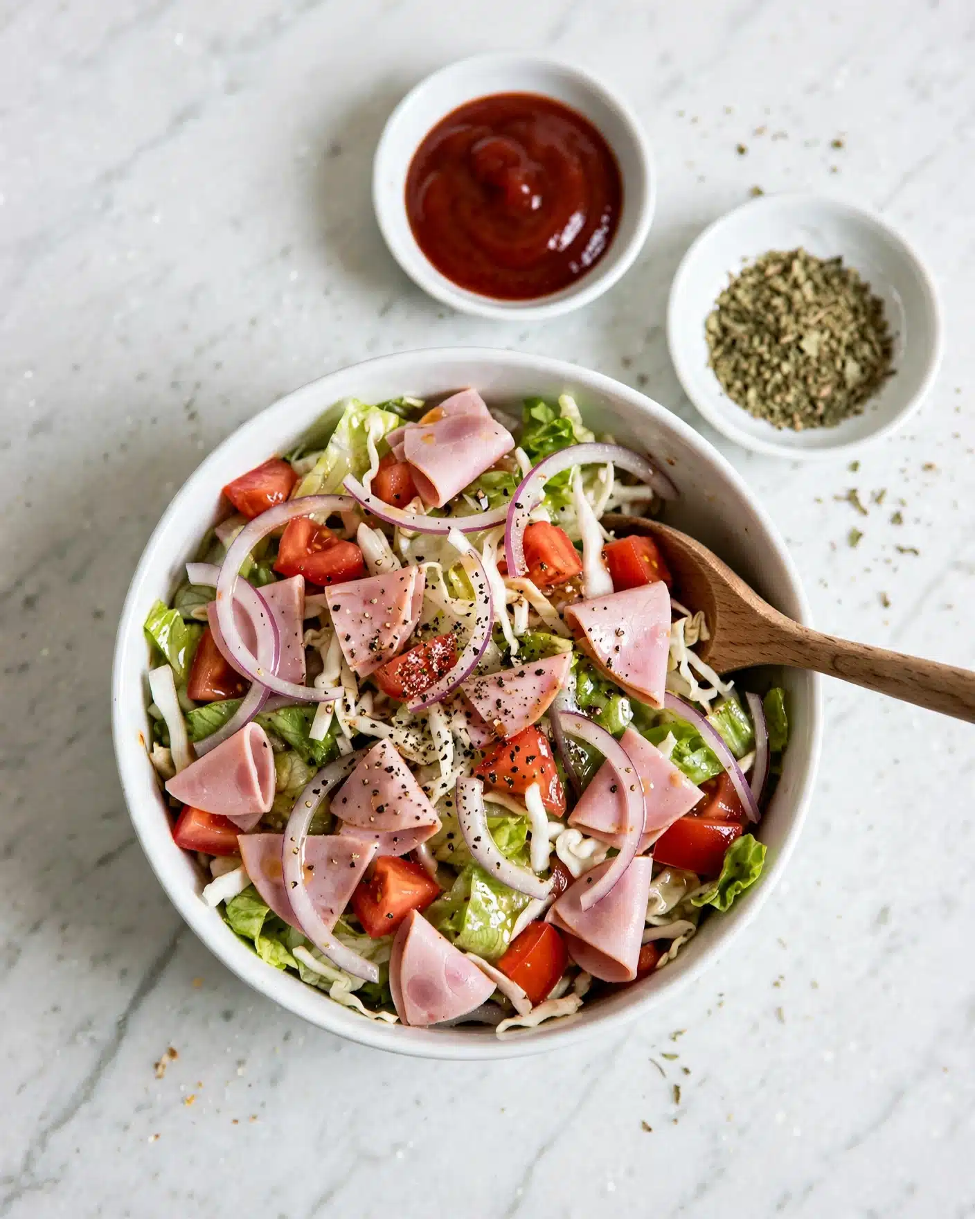 Italian Grinder Salad (Chopped Sub Salad) Recipe 5 The image shows a fresh salad inside a white bowl, filled with multiple colorful layers. The bottom layer is light green chopped lettuce mixed with pieces of red tomatoes, thin slices of light purple onion, and bits of white cabbage. There are small slices of pink deli meat mixed evenly throughout. The salad is topped with a sprinkle of coarse black pepper and is slightly glossy with a light dressing. A wooden spoon rests inside the bowl on the right side. In the background, two small white dishes hold a dark red dipping sauce and some crushed green herbs, all set on a white marbled surface. Photo taken with an iphone --ar 4:5 --v 7