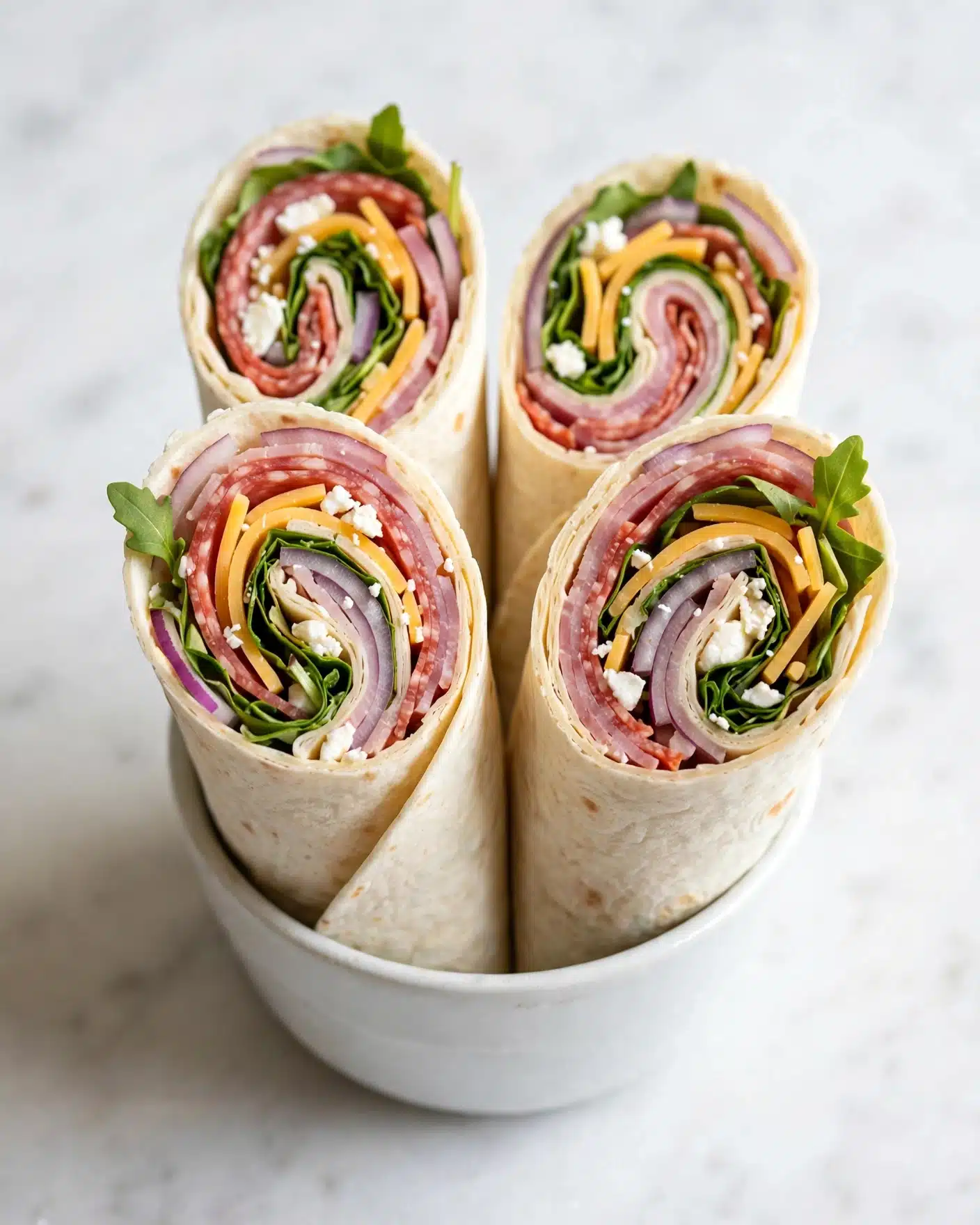 Easy Keto Salami and Italian Dressing Wraps Recipe 5 The image shows four rolled wraps placed upright inside a small white bowl on a white marbled surface. Each wrap has several layers: the outer layer is a pale cream-colored tortilla, inside it there is a reddish-pink meat layer, followed by fresh green leafy spinach and thin strips of yellow cheese with some small bits of white cheese and thinly sliced onions. The layers create a colorful spiral view from the top, showing the green, yellow, and pink colors brightly. The background is softly blurred white marbled texture, focusing attention on the wraps. photo taken with an iphone --ar 4:5 --v 7