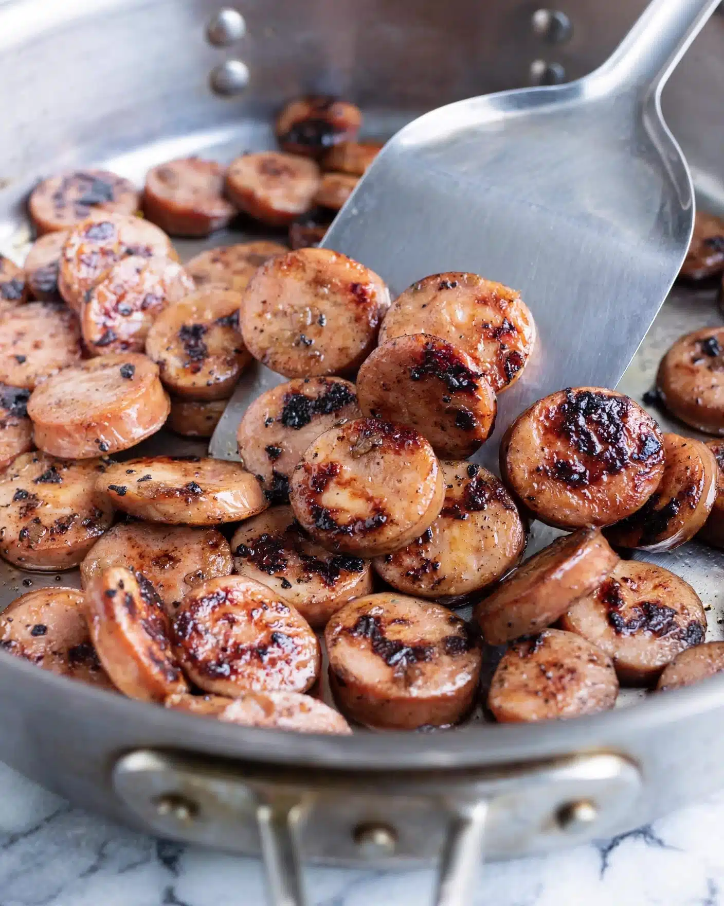 Sweet Potato and Sausage Quinoa Bowl Recipe 4 The image shows a close-up of many small round slices of sausage cooking in a metal pan. The sausages have a light brown color with dark brown and black char marks on the top, giving a grilled look. A metal spatula is lifting some slices from the pan, with the sausages layered close together. The background is a white marbled texture. photo taken with an iphone --ar 4:5 --v 7