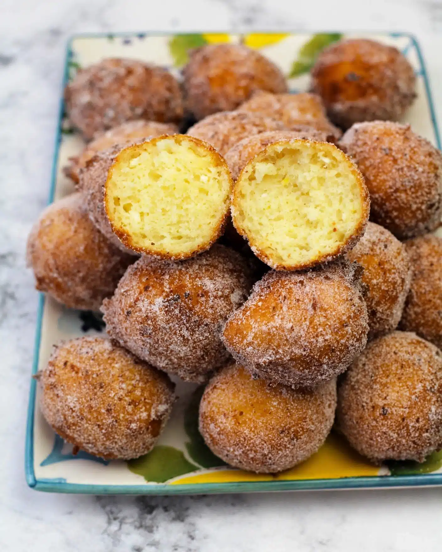 Easy Italian Lemon Ricotta Doughnuts Recipe 5 A pile of round, golden-brown doughnuts coated in a fine layer of sugar covers a white plate with colorful edges. Two doughnuts sit on top, split open to show their soft, airy, pale yellow inside with a light, fluffy texture. The sugar on the outside looks slightly grainy and evenly coats the doughnuts. The plate is set on a white marbled surface. photo taken with an iphone --ar 4:5 --v 7