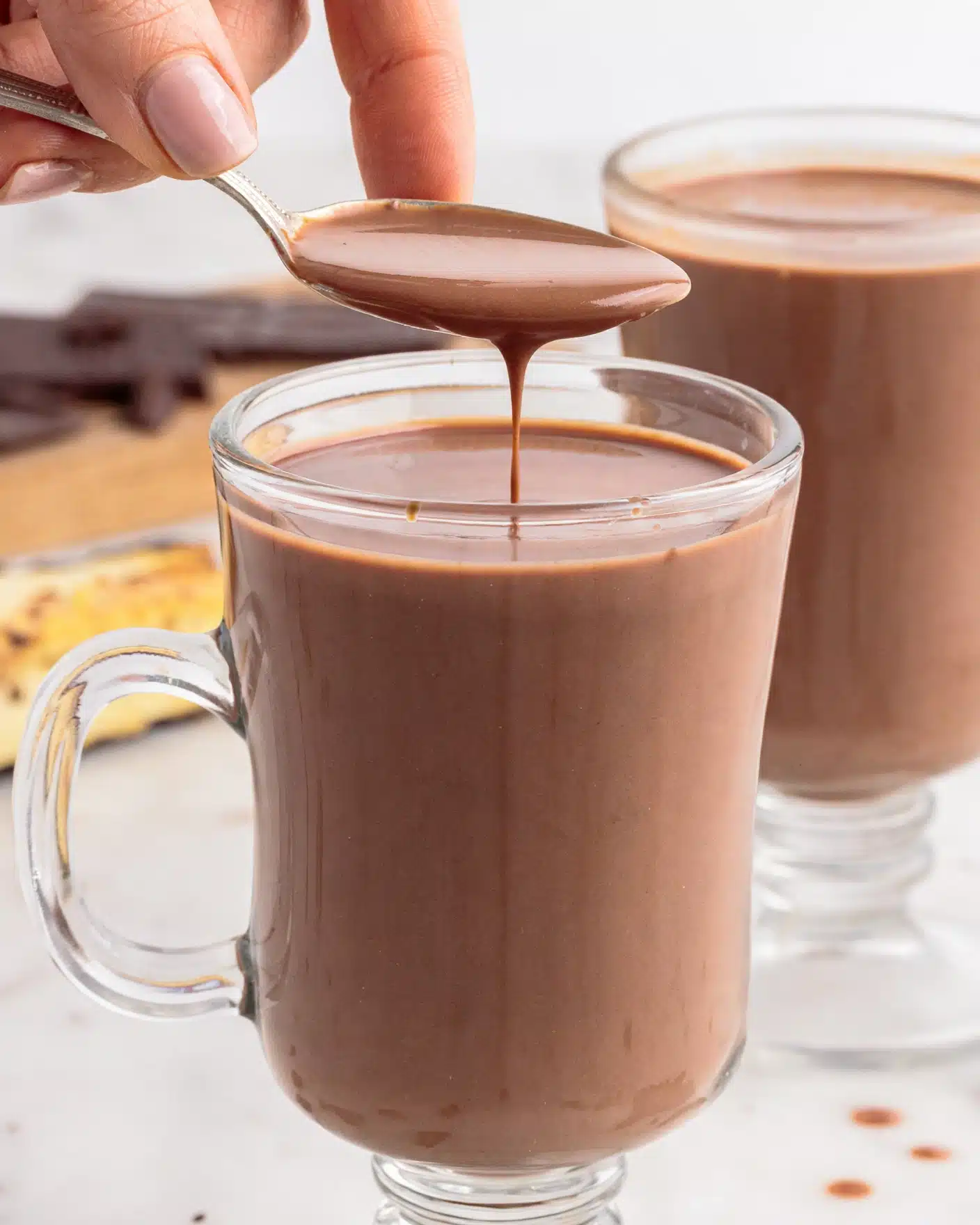 The image shows two clear glass mugs filled with smooth, rich chocolate drink, placed on a white marbled surface. In the front mug, a spoon is held by a woman's hand above it, dripping thick chocolate liquid back into the mug. The chocolate drink has a shiny, silky texture and a deep brown color. The background is softly blurred with pieces of dark chocolate and a white object with yellow and brown patterns. The photo captures the smooth drip of chocolate in midair, focusing on the glass and spoon with natural light. photo taken with an iphone --ar 4:5 --v 7