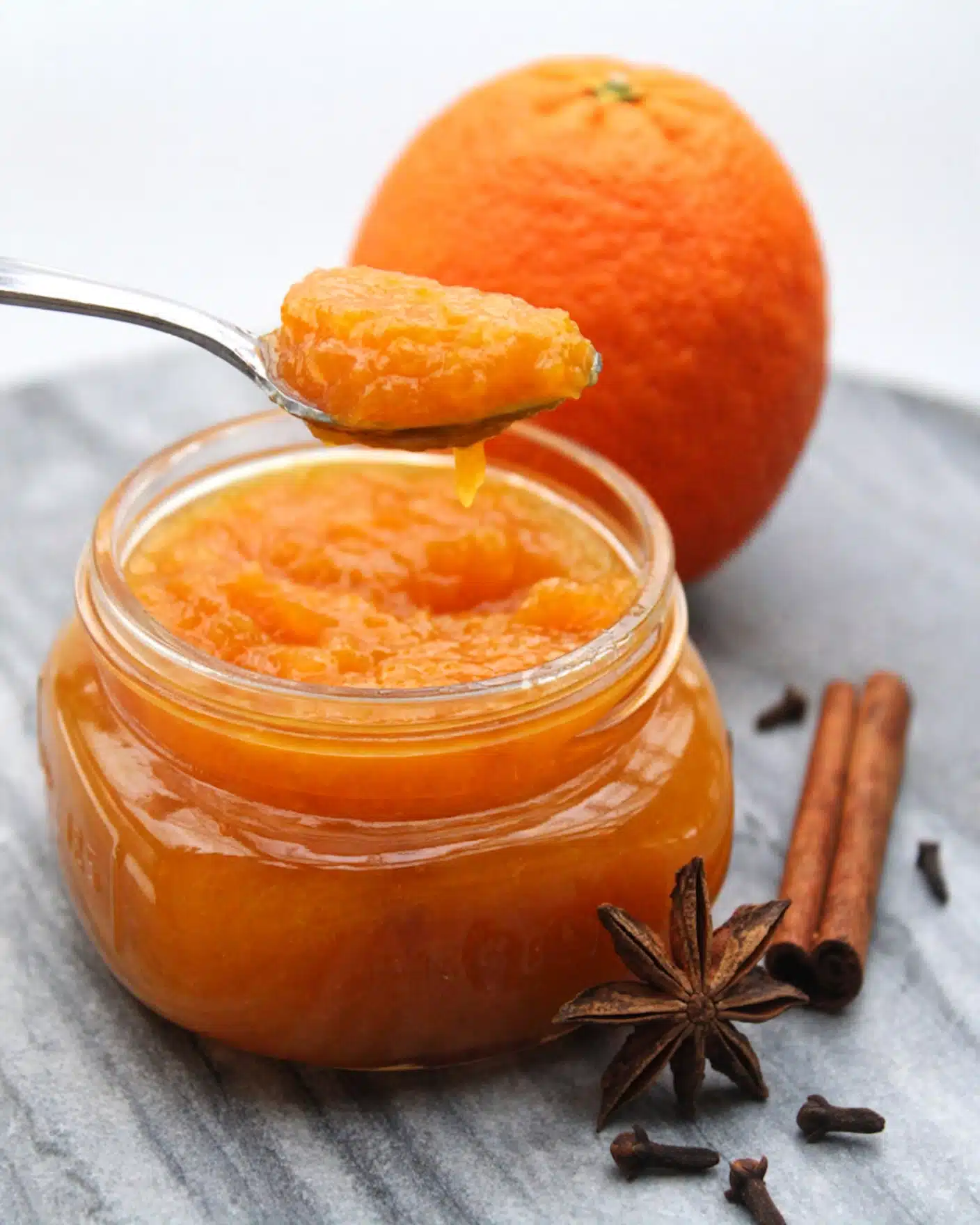 Orange Jam Recipe 5 A small clear glass jar filled with bright orange jam with a thick, smooth texture, shown close up with a shiny silver spoon holding a scoop of jam above the jar. Behind the jar is a whole, textured orange fruit with a vibrant orange color. Next to the jar on the white marbled surface are two cinnamon sticks that are brown and rough, along with a dark brown star anise and a few scattered small cloves. The scene is softly lit, focusing on the rich colors and textures. Photo taken with an iphone --ar 4:5 --v 7