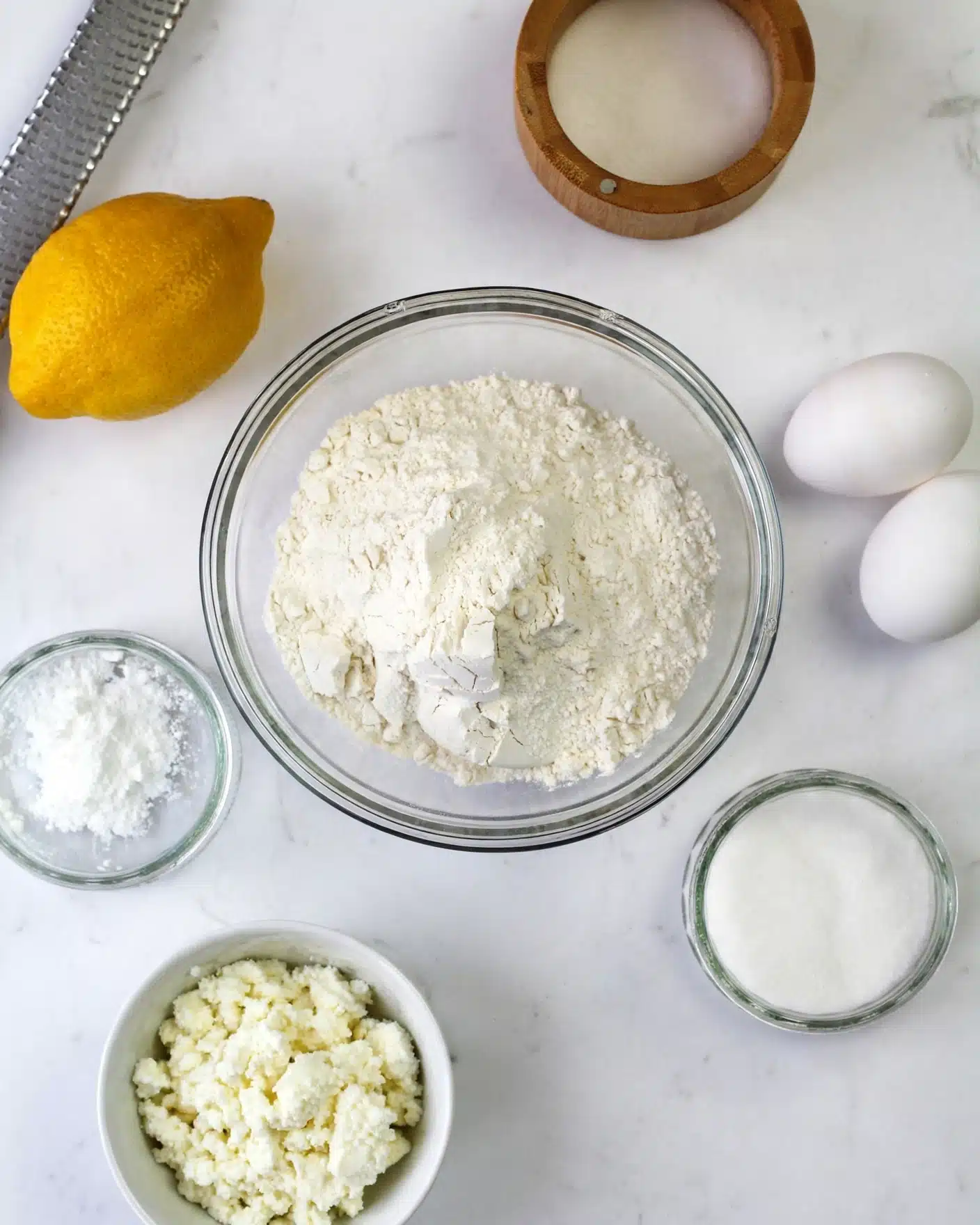 Easy Italian Lemon Ricotta Doughnuts Recipe 4 A clear glass bowl filled with a mound of white flour sits in the center on a white marbled surface. Surrounding it are several small containers: a wooden bowl with white sugar at the top right, a small clear glass bowl with white granulated sugar at the bottom right, and a small clear glass bowl with white powder, possibly baking powder, to the left. A bright yellow lemon is placed toward the top left. Additionally, two white eggs rest on the white marbled surface to the right, and a white bowl holds a soft, crumbled white cheese at the bottom left. A large metal grater is partially visible in the top left corner of the image. photo taken with an iphone --ar 4:5 --v 7