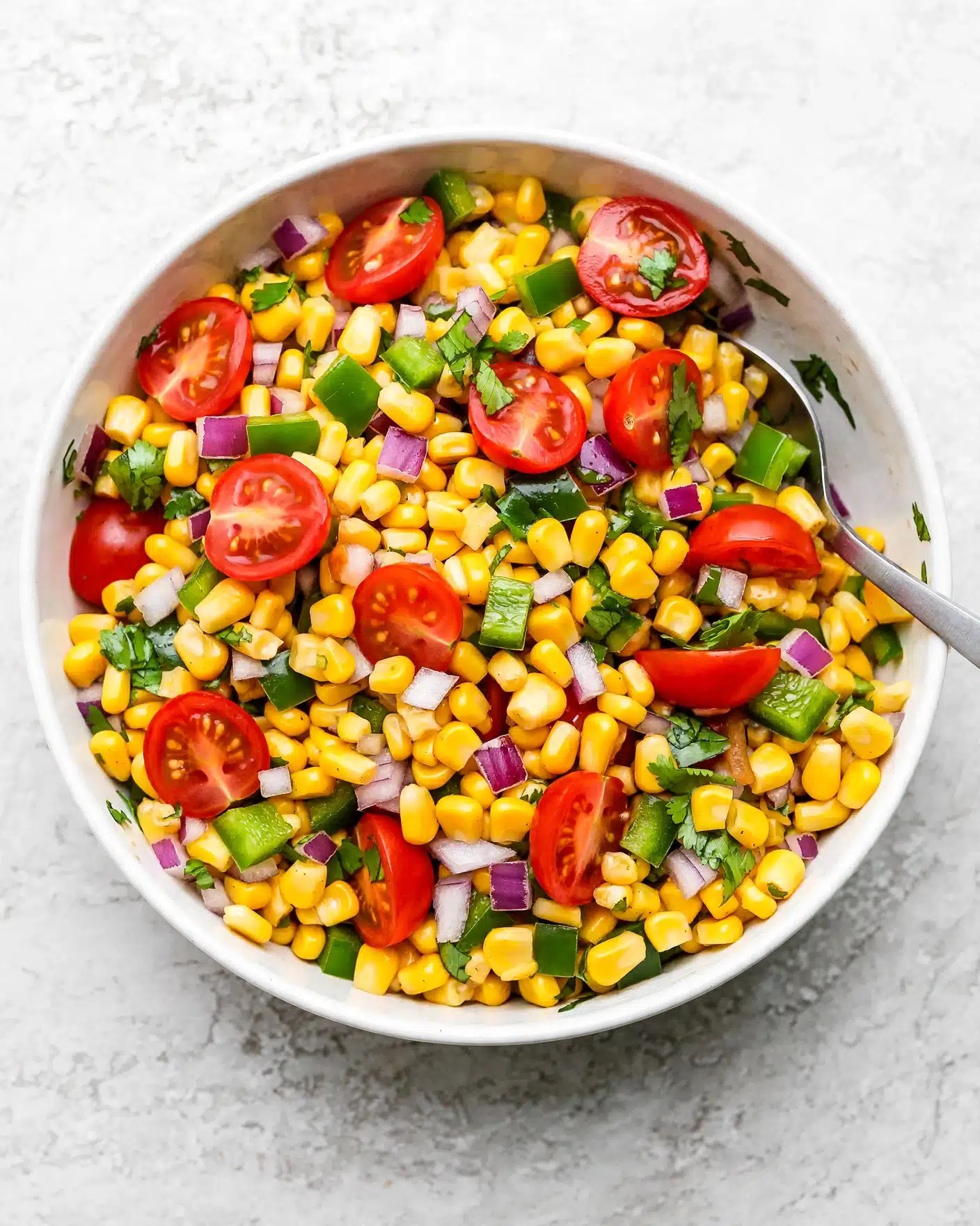 Steak Fajita Bowl Recipe 4 A close-up view of a white bowl filled with a colorful corn salad showing four layers: the bottom layer is whole yellow corn kernels, scattered throughout are pieces of bright red cherry tomatoes cut in halves, small diced purple onions adding a rough texture, and freshly chopped green herbs including cilantro and green pepper. All ingredients are mixed together, making a fresh and vibrant look with different sizes and shapes of each vegetable. The bowl sits on a white marbled surface with a metal spoon partially visible on the right side inside the bowl. Photo taken with an iphone --ar 4:5 --v 7