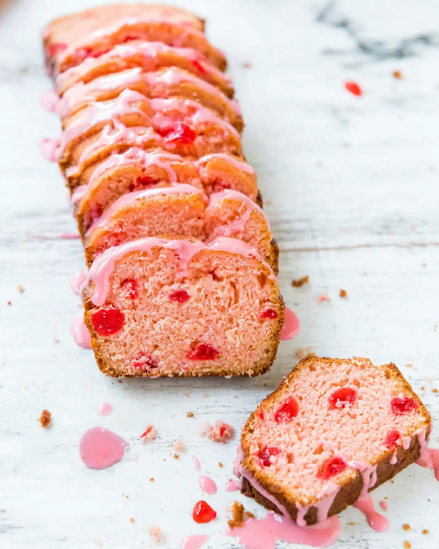 Sweet Soft Cherry Bread with Cherry-Almond Glaze Recipe 5 The image shows a loaf of sliced cake with a pink color and red fruit bits inside each slice. The cake has about eight slices arranged in a row on a white marbled surface. Each slice is topped with a light pink glaze that drips down the sides. Crumbs and drops of glaze are scattered around the cake, adding a casual look. The overall texture of the cake looks soft and moist. photo taken with an iphone --ar 4:5 --v 7
