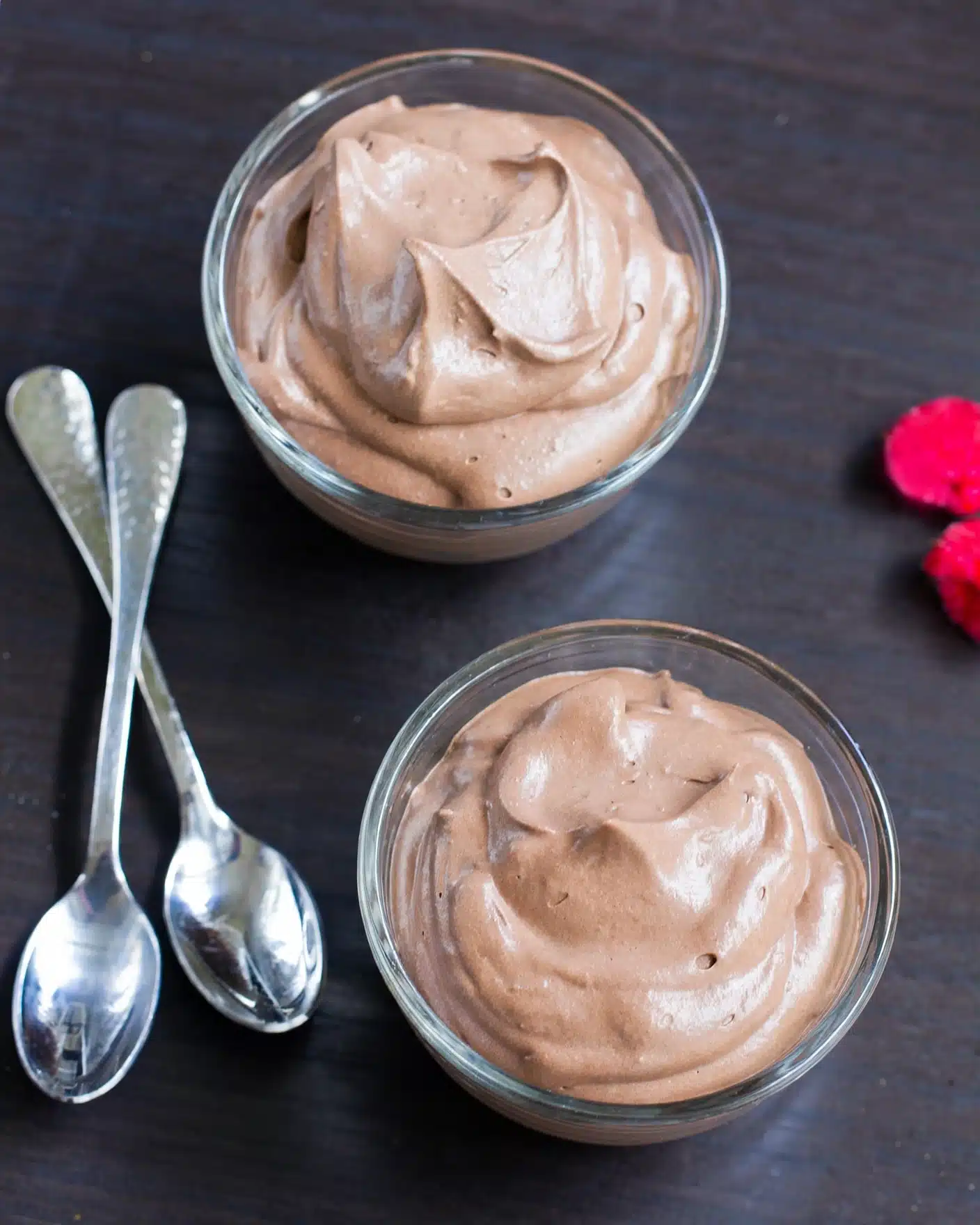 Two clear glass bowls each hold a single layer of smooth, creamy chocolate mousse with soft peaks on top, showing a light and fluffy texture. The mousse is rich brown and fills each bowl nearly to the rim. The bowls sit on a dark wooden surface, beside two shiny silver spoons arranged neatly next to each other. In the corner, there is a small red flower adding a bright color contrast. photo taken with an iphone --ar 4:5 --v 7