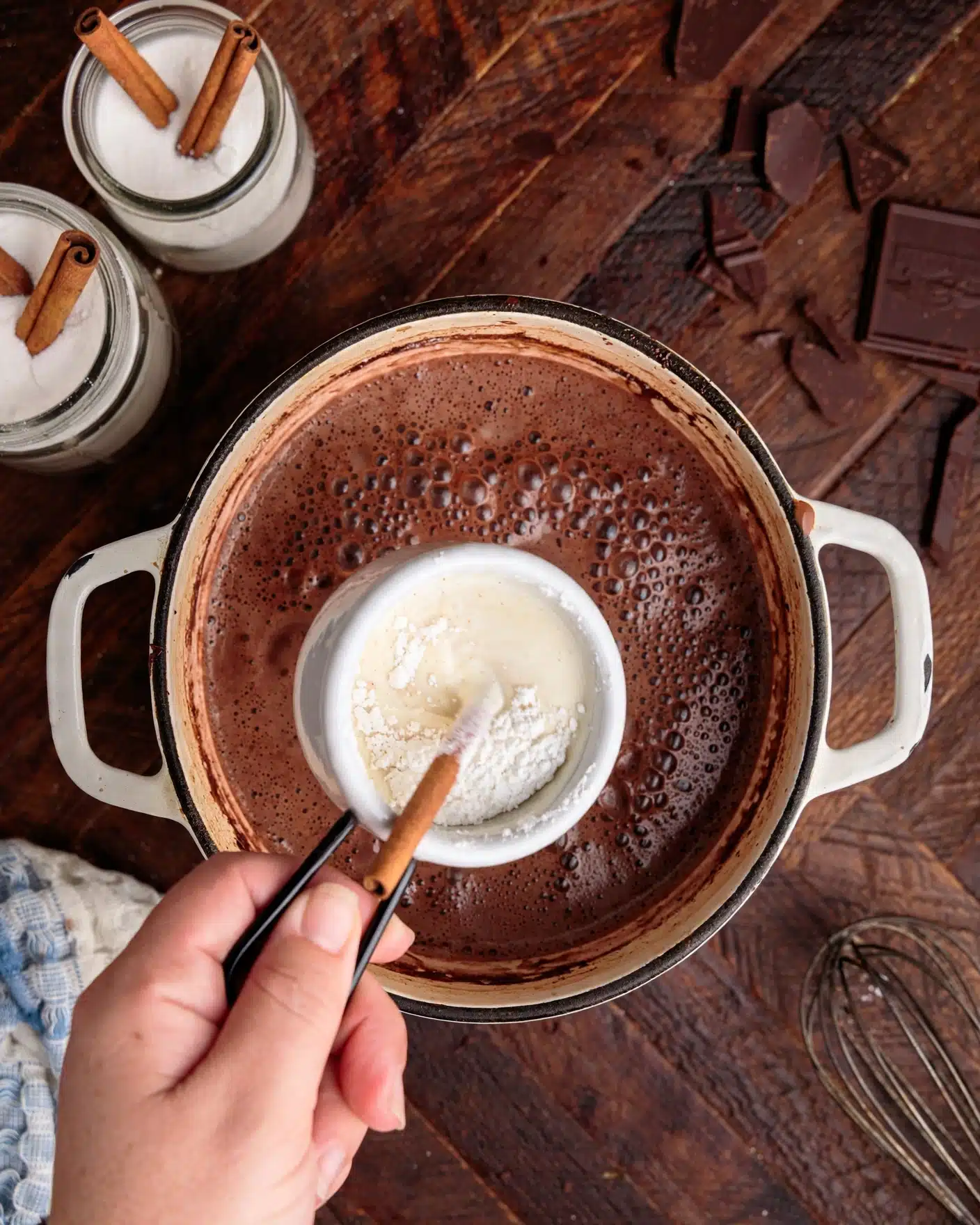 A white pot filled with frothy dark brown hot chocolate base showing bubbles and light brown rim stains holds a smaller white measuring cup over it, which contains white powder mixed with a pale cream liquid stirred by a light brown stick. A woman's hand grips the black handle of the measuring cup gently. Nearby on a dark wood surface are pieces of broken dark chocolate, two glass jars with white contents topped with cinnamon sticks, and a metal whisk. The scene is cozy and warm, photo taken with an iphone --ar 4:5 --v 7