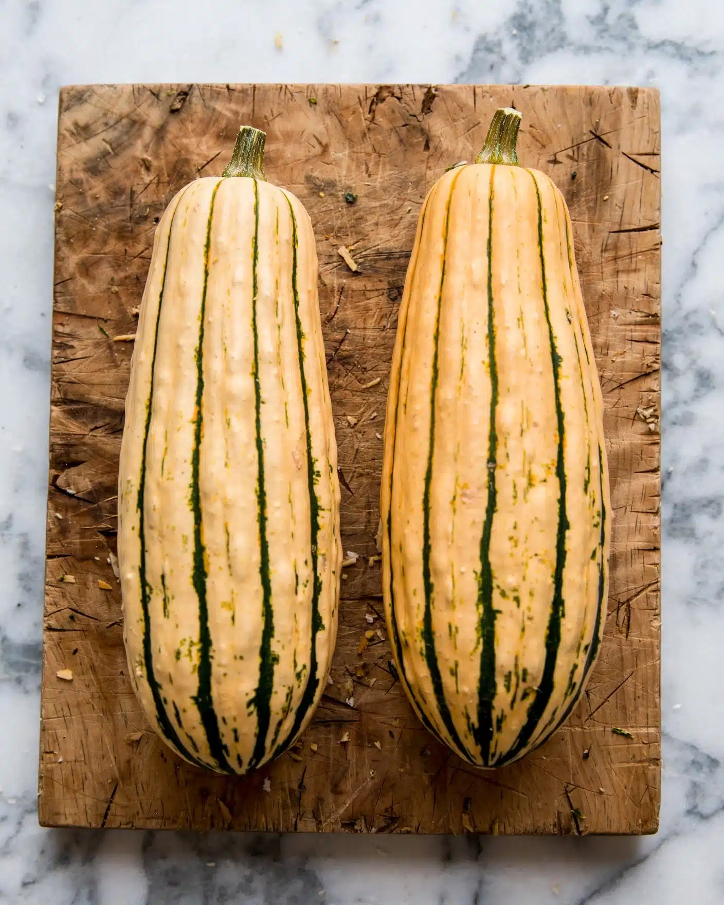 The image shows two whole delicata squash placed side by side on a wooden cutting board. Both squash are elongated with a smooth texture and light orange skin featuring dark green vertical stripes running from top to bottom. The tops of each squash have short, sturdy stems. The cutting board has a natural wood grain with visible knife marks and sits on a white marbled surface. Photo taken with an iphone --ar 4:5 --v 7