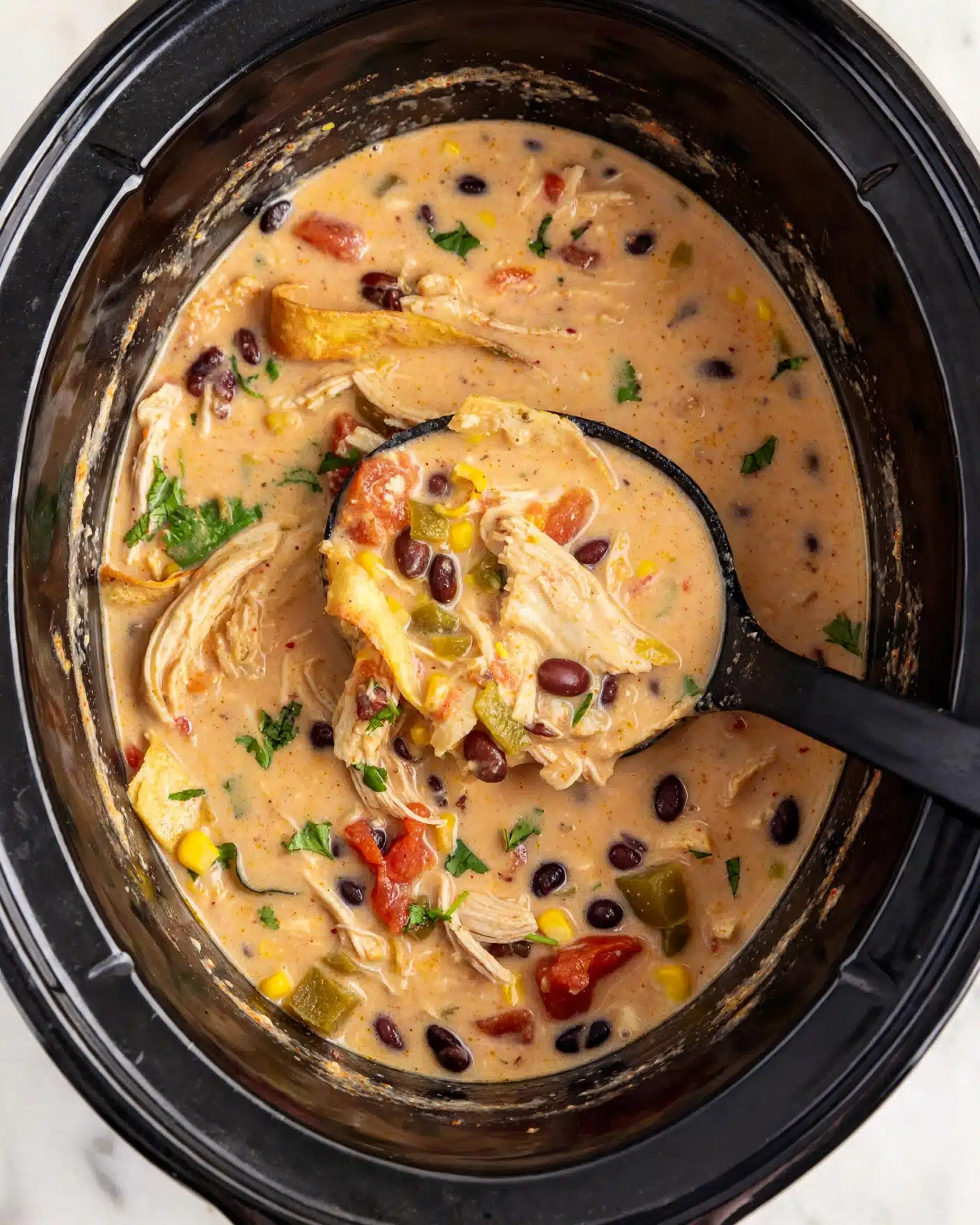 Creamy Chicken Tortilla Soup in the Crock Pot Recipe 5 The image shows a close-up of a creamy soup with several visible layers and ingredients. The base layer is a thick, light orange broth with a smooth texture, filling the entire white marbled surface. Within the soup, there are large pieces of shredded light-colored chicken placed unevenly throughout. Scattered among the chicken are black beans, small yellow corn kernels, and chunks of red tomatoes, adding pops of dark brown, yellow, and red color. Small green pieces, likely cilantro or herbs, are sprinkled on top for freshness. A black ladle is scooping up a portion of the soup, showing these ingredients clearly inside the ladle bowl. The overall scene is warm and cozy, with a focus on the rich mixture of textures and colors. Photo taken with an iphone --ar 4:5 --v 7