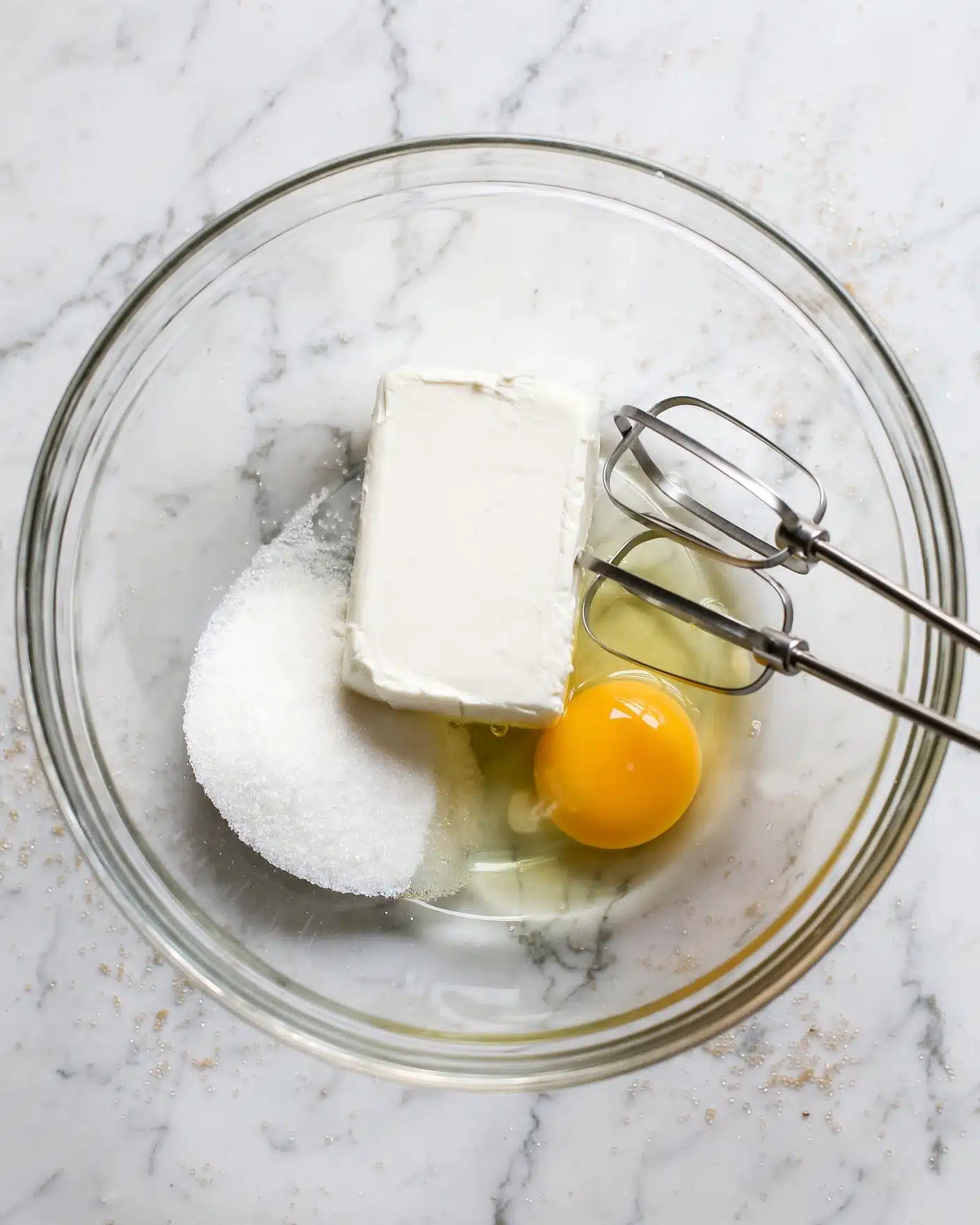 Strawberry Cheesecake Dump Cake Recipe 3 A clear glass bowl sits on a white marbled surface, holding three main ingredients: a white block of cream cheese in the center, a pile of white sugar to the left, and a single raw egg with a bright yellow yolk to the right. Two silver metal beaters rest inside the bowl on the right side, ready to mix the ingredients. The scene is simple and clean with soft natural light. Photo taken with an iphone --ar 4:5 --v 7