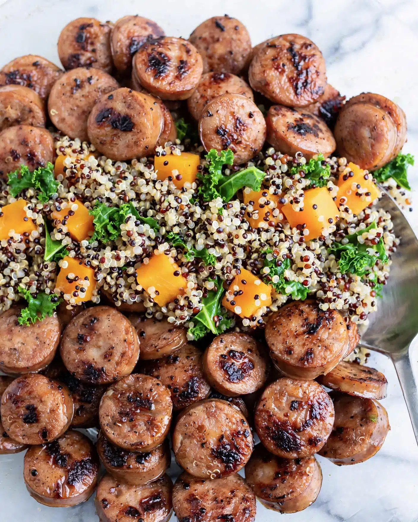 Sweet Potato and Sausage Quinoa Bowl Recipe 5 A close-up of a dish with three main layers: small round slices of browned sausage on the bottom and top, soft quinoa in black, white, and light brown colors filling most of the middle, and scattered bright orange cubes of butternut squash along with dark green kale leaves mixed evenly across the dish, all seen on a white marbled surface with a metal spoon partially visible on the right side of the image photo taken with an iphone --ar 4:5 --v 7