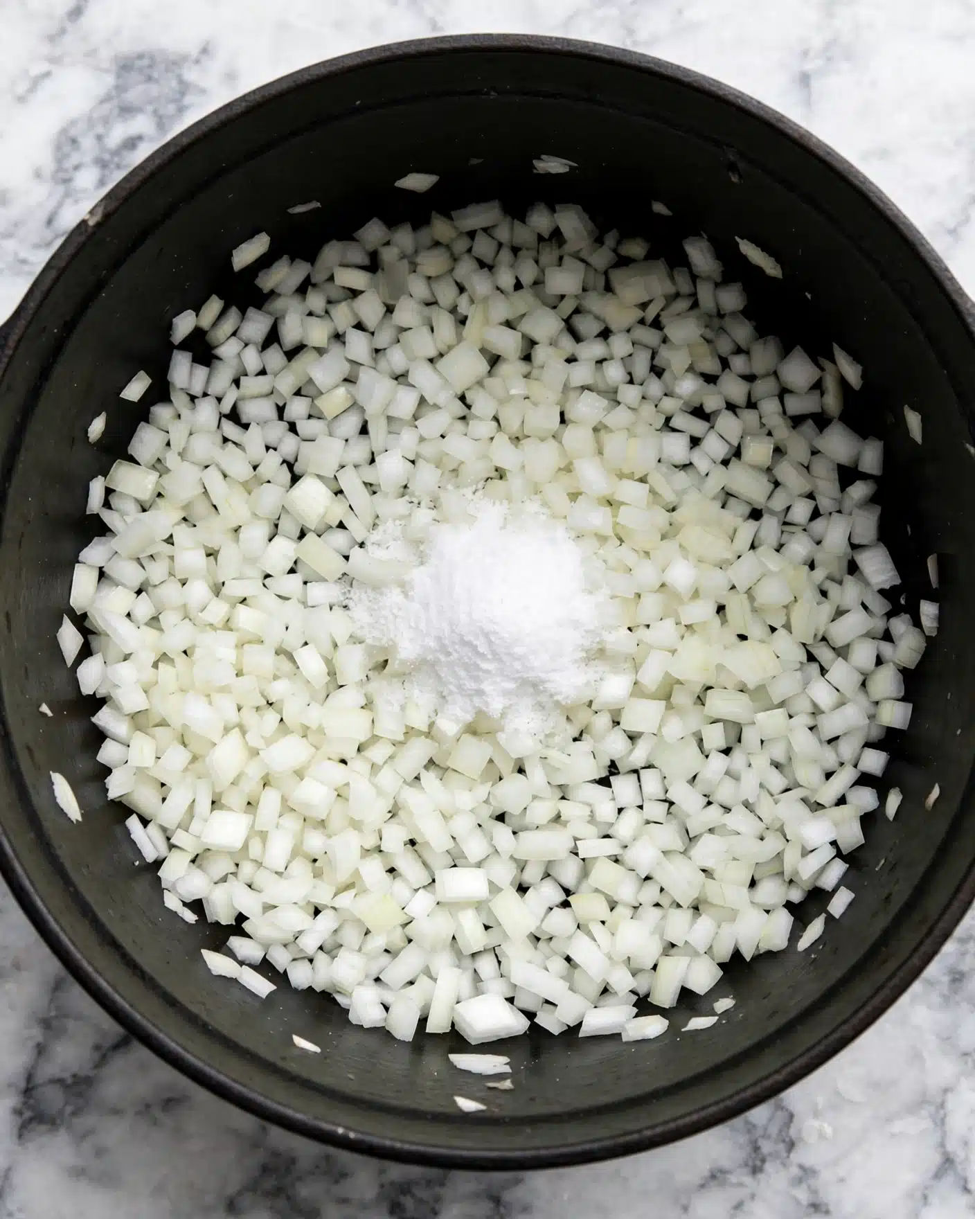 A close-up image shows a black pot filled with many small white onion pieces spread evenly inside. In the center, there is a small pile of white granulated salt resting on top of the chopped onions. The pot is sitting on a white marbled surface. Photo taken with an iphone --ar 4:5 --v 7