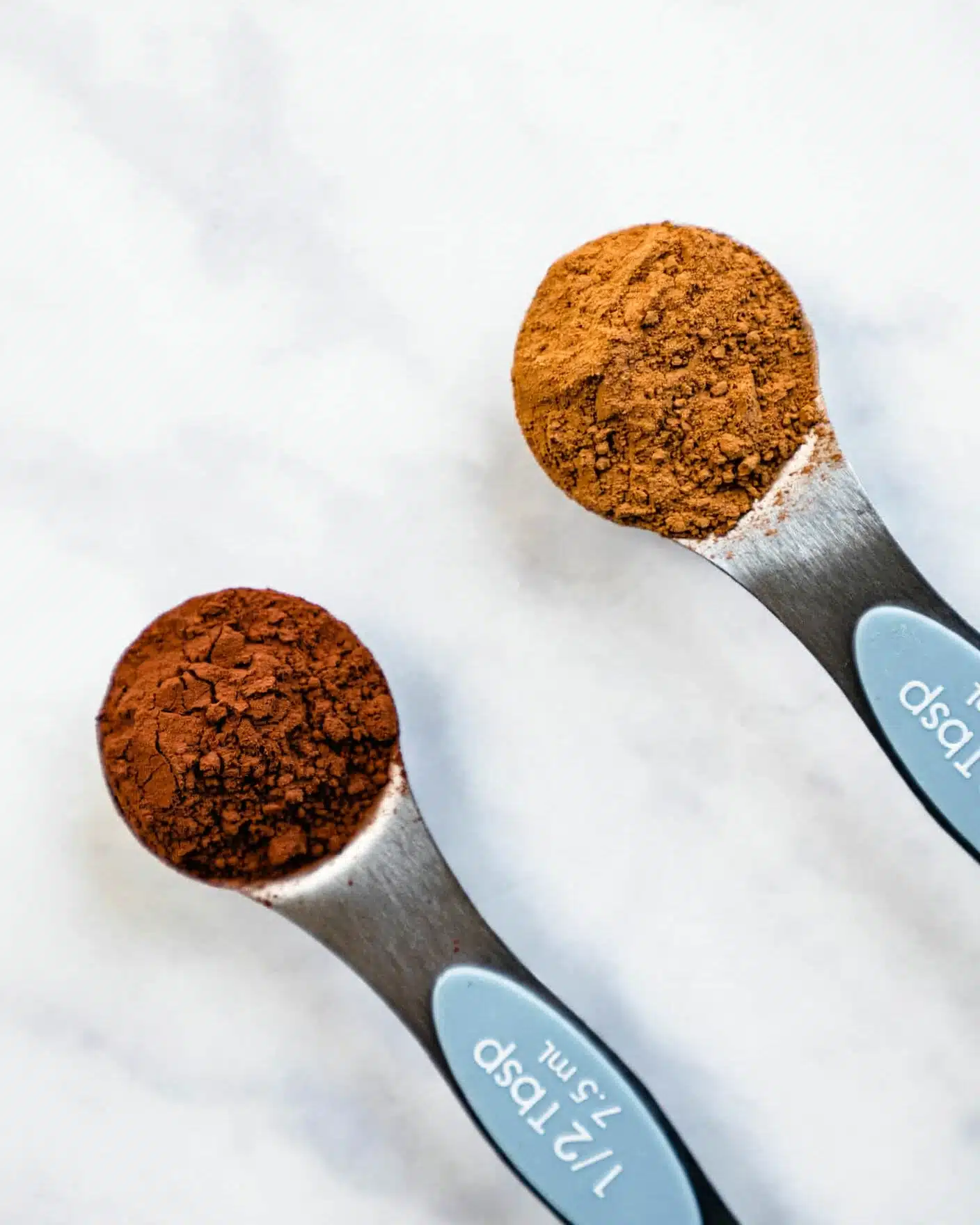 Two metal measuring spoons rest on a white marbled surface, each filled with a different brown powder. The left spoon holds a darker brown, almost reddish powder with a rough, crumbly texture, while the right spoon contains a lighter, warm brown powder that appears finer and smoother. The spoons have soft blue handles with white text showing measurements. The light softly highlights the texture and colors of the powders. photo taken with an iphone --ar 4:5 --v 7