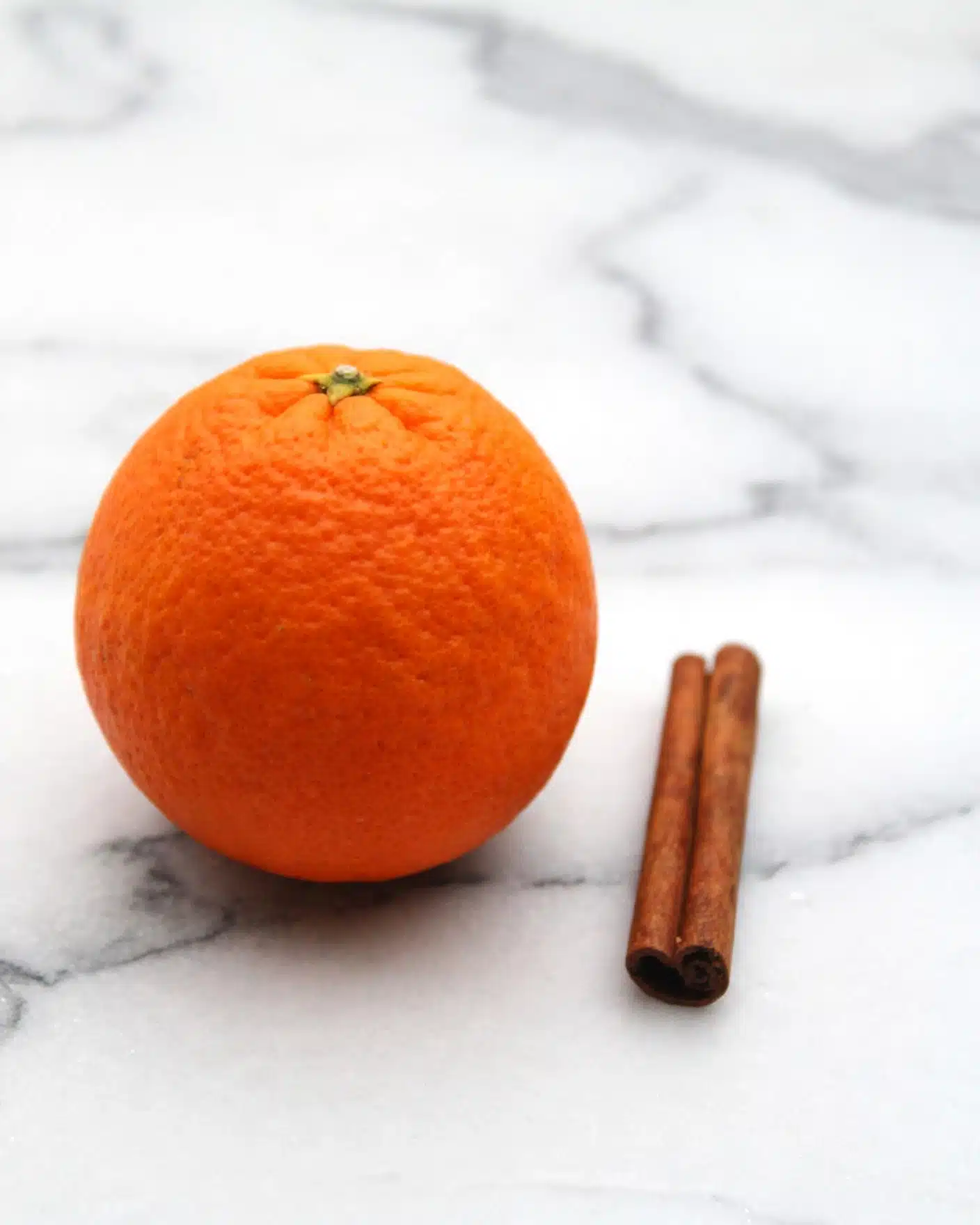Orange Jam Recipe 4 The image shows a bright orange placed on a white marble surface with light gray veins. The orange has a bumpy, textured skin that is evenly colored and round in shape. Next to the orange are two cinnamon sticks, dark brown with a rough and slightly curved texture, lying parallel to each other on the marble. The overall composition is simple, with the orange as the main focus and the cinnamon sticks adding contrast. photo taken with an iphone --ar 4:5 --v 7