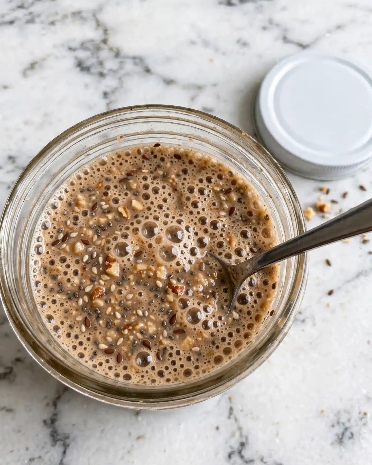 Brown Sugar Shaken Espresso Overnight Oats Recipe 4 A clear glass jar filled with a bubbly, light brown sauce containing small bits of nuts and seeds is shown from above. A metal spoon is dipped inside the jar next to the bubbles on the sauce surface. The jar’s white lid is placed on the white marbled surface nearby. photo taken with an iphone --ar 4:5 --v 7