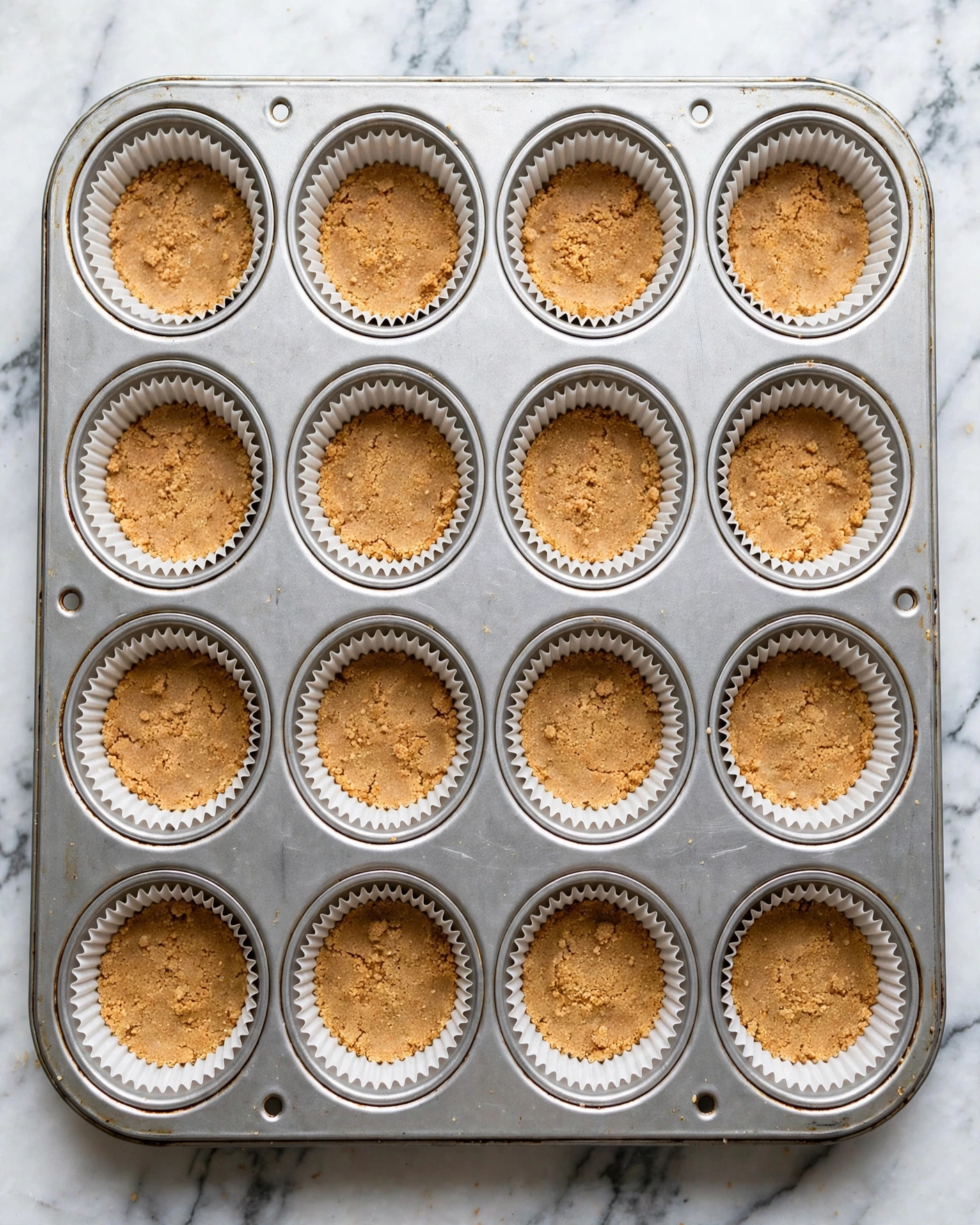 Mini Mango Lassi Cheesecakes (No-Bake) Recipe 4 A silver metal muffin tray with twelve round wells is shown from a close angle. Each well contains a white paper liner holding a thin, even layer of light brown crumb mixture pressed flat against the bottom. The tray is placed on a surface with a white marbled texture. The crumb layers have a slightly grainy texture and a warm, golden-brown color, offering a clear contrast to the white liners and silver tray. photo taken with an iphone --ar 4:5 --v 7