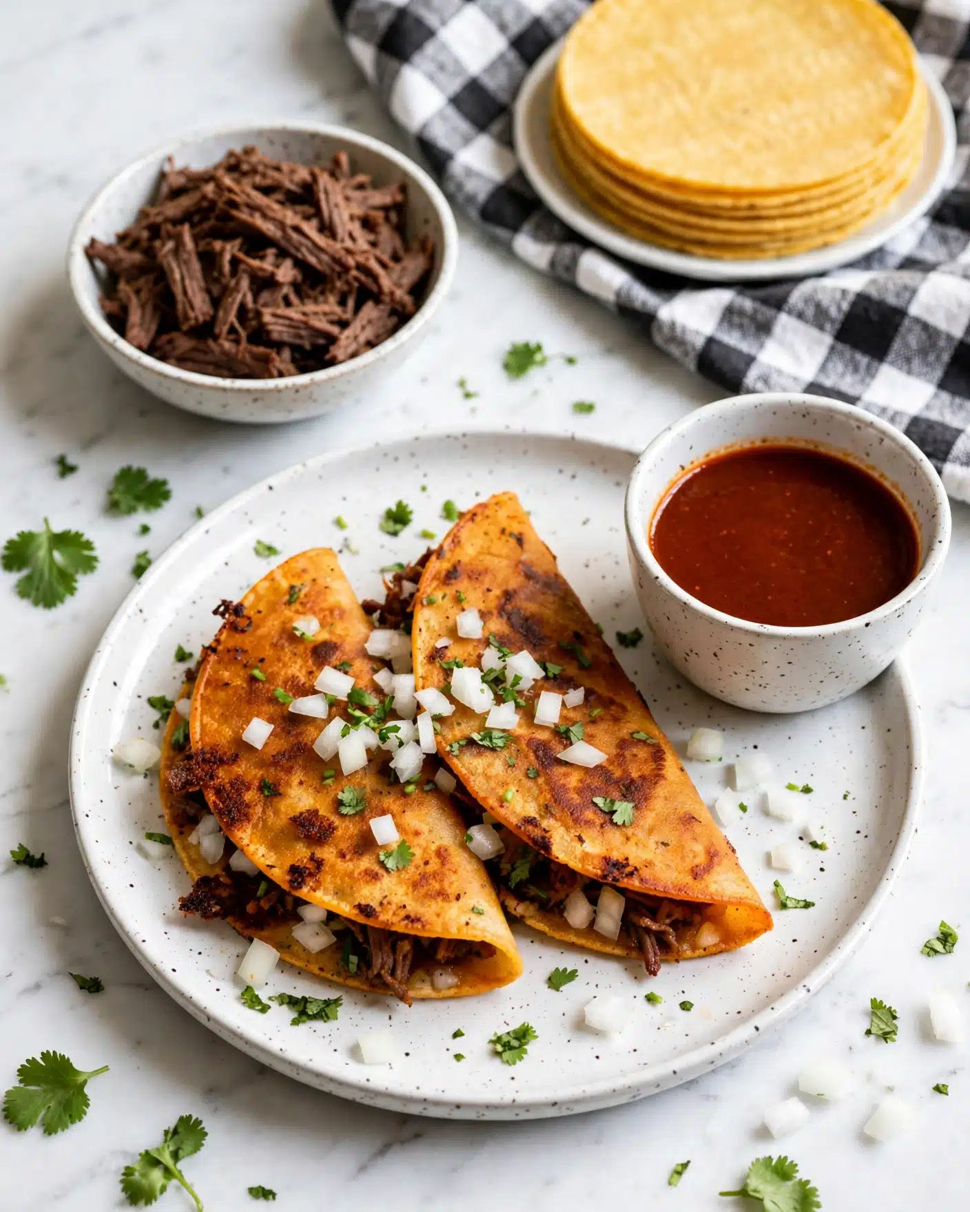 Slow Cooker Birria Tacos Recipe 5 Two golden-brown folded tacos lie on a white speckled plate, each taco sprinkled with small bits of white onion and chopped green cilantro. The tacos have a slightly crispy texture with some darker spots showing where they are fried. Near the tacos is a small, round white speckled cup filled with deep red sauce. In the background on the white marbled surface, a bowl filled with finely shredded dark brown meat is visible, as well as a white dish holding stacked yellow tortillas resting on a black and white checkered cloth. Scattered cilantro leaves and onion pieces add a fresh touch around the plate. Photo taken with an iphone --ar 4:5 --v 7
