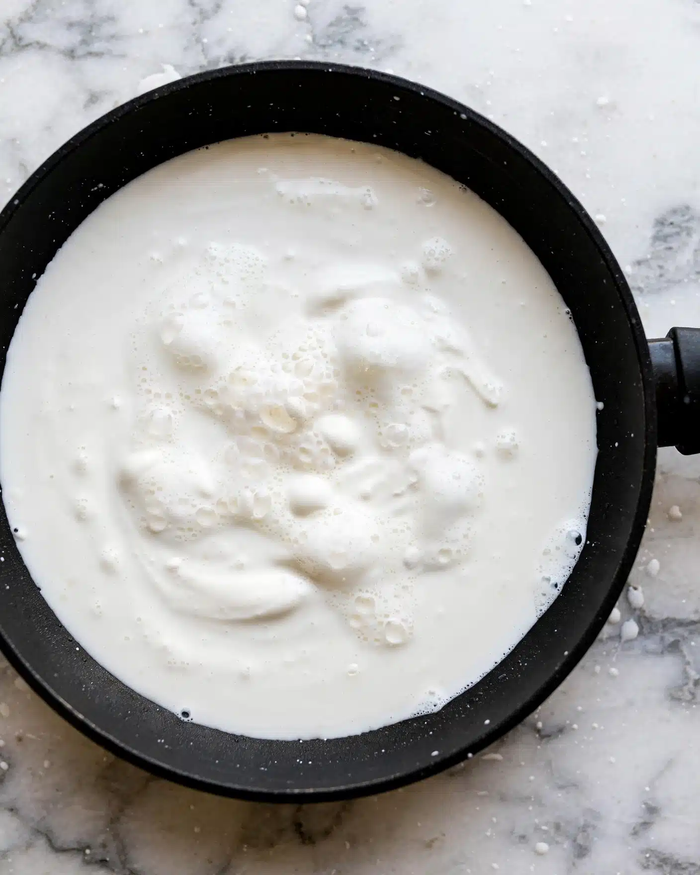 A close-up top view of a black pan filled with smooth white liquid cream, with some small bubbles and foam in the center, placed on a white marbled surface. The pan's handle is partially visible on the right side. photo taken with an iphone --ar 4:5 --v 7
