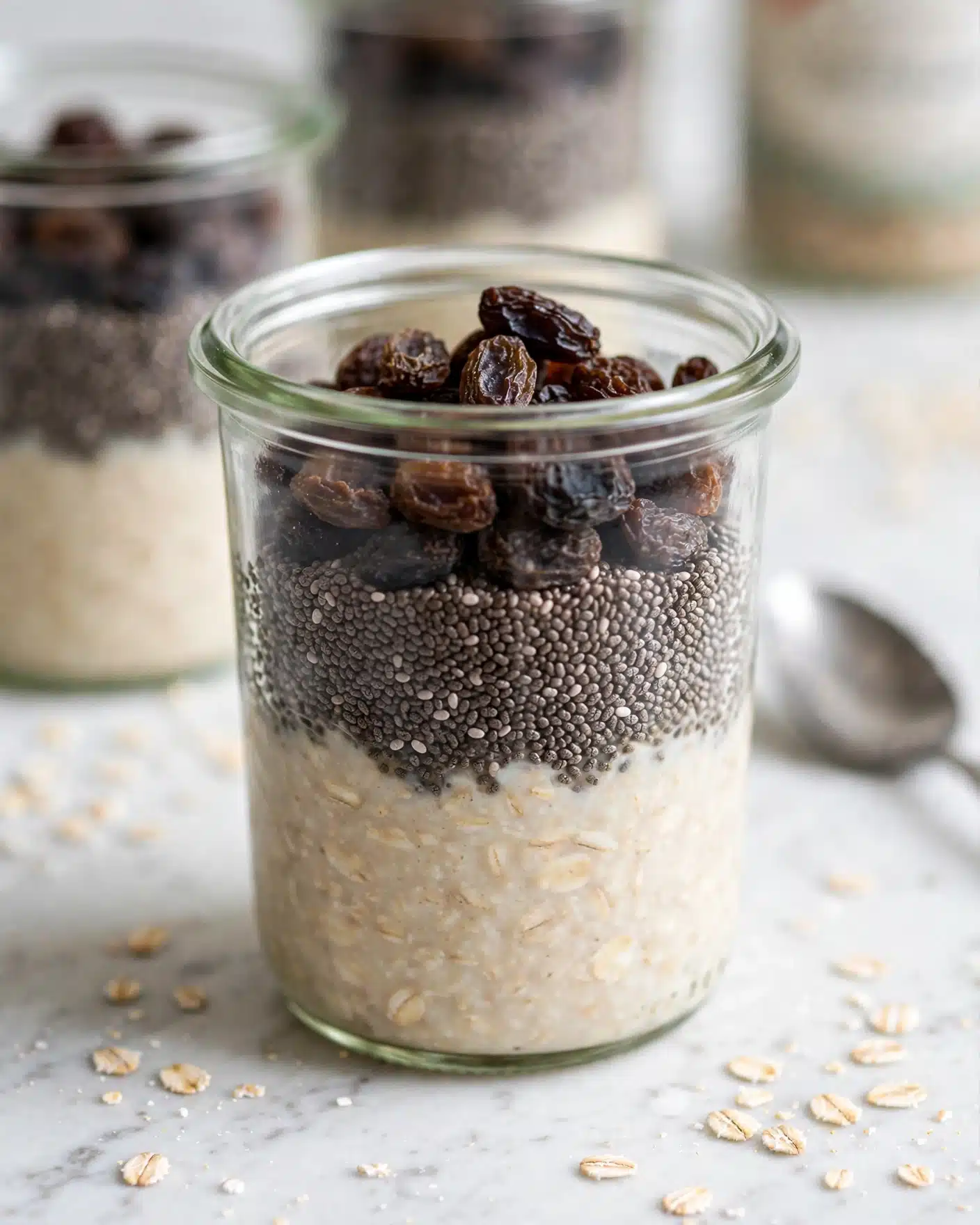 The image shows a clear glass jar filled with three distinct layers resting on a white marbled surface. The bottom layer is a thick, creamy oatmeal with a pale beige color and slightly textured surface. Above that is a middle layer of small, dark chia seeds tightly packed, adding a rough texture and deep blackish-brown shade. The top layer consists of dark raisins scattered unevenly, showing a wrinkled texture and a mix of dark brown and black tones. In the blurred background, another similar jar is visible, along with a silver spoon. Scattered oatmeal flakes add a light touch to the white marbled surface. photo taken with an iphone --ar 4:5 --v 7