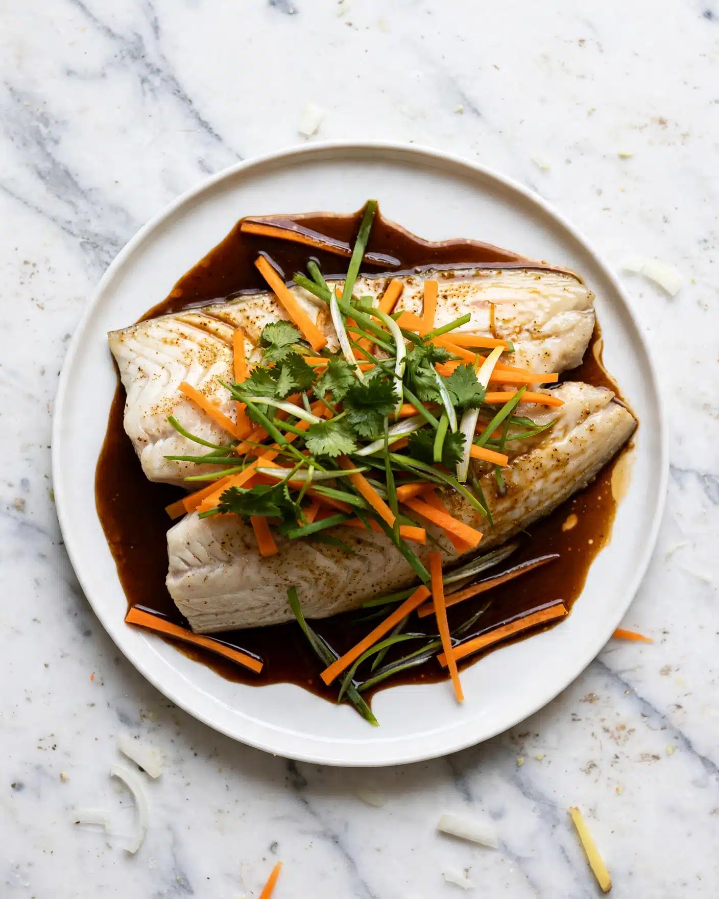 A white plate with a thick white fish fillet as the bottom layer, slightly shiny with light brown seasoning and sauce around it. On top of the fish, a mix of thin carrot sticks, green onion strips, and fresh green cilantro leaves are stacked in a loose pile, giving bright orange, light green, and dark green colors. The sauce pools around the fish, dark brown and glossy, with some thin green vegetable strips lying in it. The background is a white marbled texture. Photo taken with an iphone --ar 4:5 --v 7