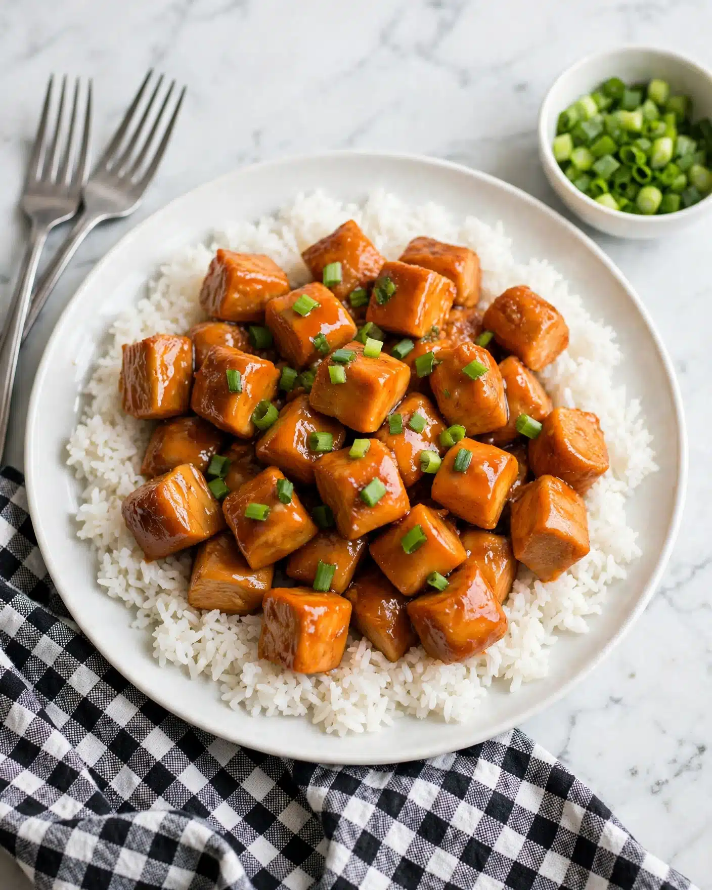 Baked Sweet and Sour Chicken Recipe 5 A white plate filled with a base layer of fluffy white rice, evenly spread to cover the entire plate. On top, there is a thick layer of bite-sized pieces of chicken coated in a rich, shiny orange sauce, giving a glossy texture. Scattered over the chicken are small pieces of bright green chopped scallions adding a fresh contrast. Next to the plate on a white marbled surface, a small white bowl holds more chopped green scallions. A black and white checked cloth is partially visible under the plate. Also, two silver forks lie beside the plate. Photo taken with an iphone --ar 4:5 --v 7