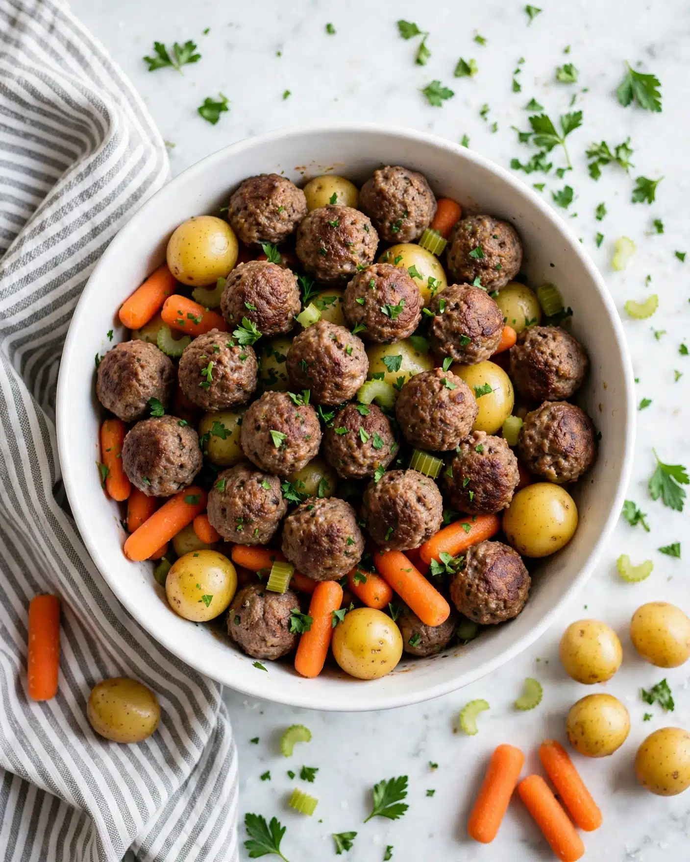 Meatball Pot Roast in the Slow Cooker Recipe 5 A white bowl filled with layers of small round meatballs that are brown and textured, baby yellow potatoes that have a smooth surface, and bright orange baby carrots. The vegetables and meatballs are mixed together and sprinkled with bits of green parsley and small pieces of celery. The bowl sits on a white marbled surface with scattered carrots, potatoes, and parsley leaves nearby, along with a gray and white striped cloth on the left side. photo taken with an iphone --ar 4:5 --v 7