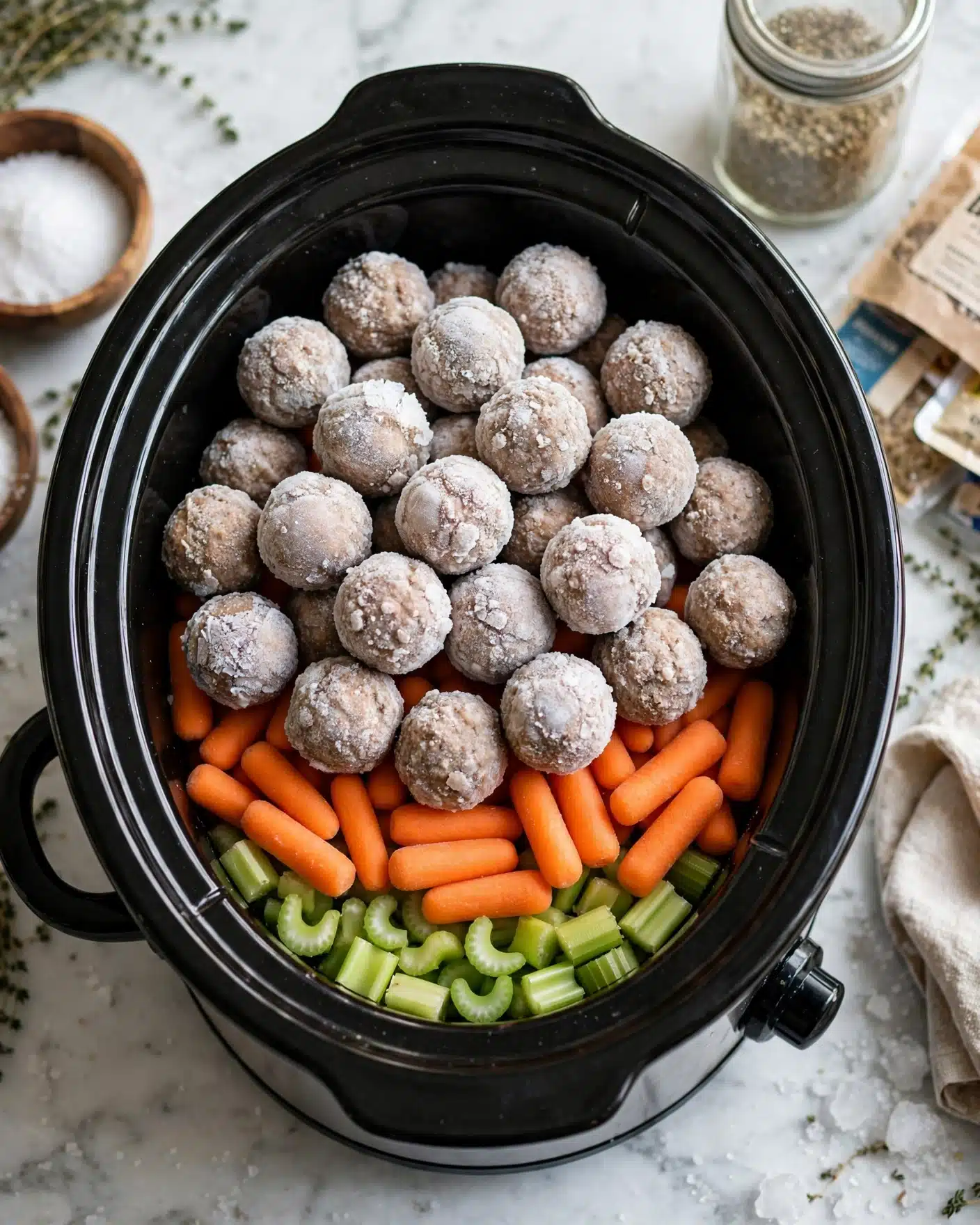 Meatball Pot Roast in the Slow Cooker Recipe 4 The image shows a black slow cooker filled with three visible layers. The bottom layer contains chopped green celery sticks arranged evenly. Above the celery is a layer of small orange baby carrots, spread out to cover the celery. The top layer consists of numerous round, frozen meatballs coated with a light white frost, piled to cover the vegetables completely. The slow cooker is placed on a white marbled surface, with partial views of a wooden bowl of salt, a clear glass jar, and some packaged food items nearby. photo taken with an iphone --ar 4:5 --v 7