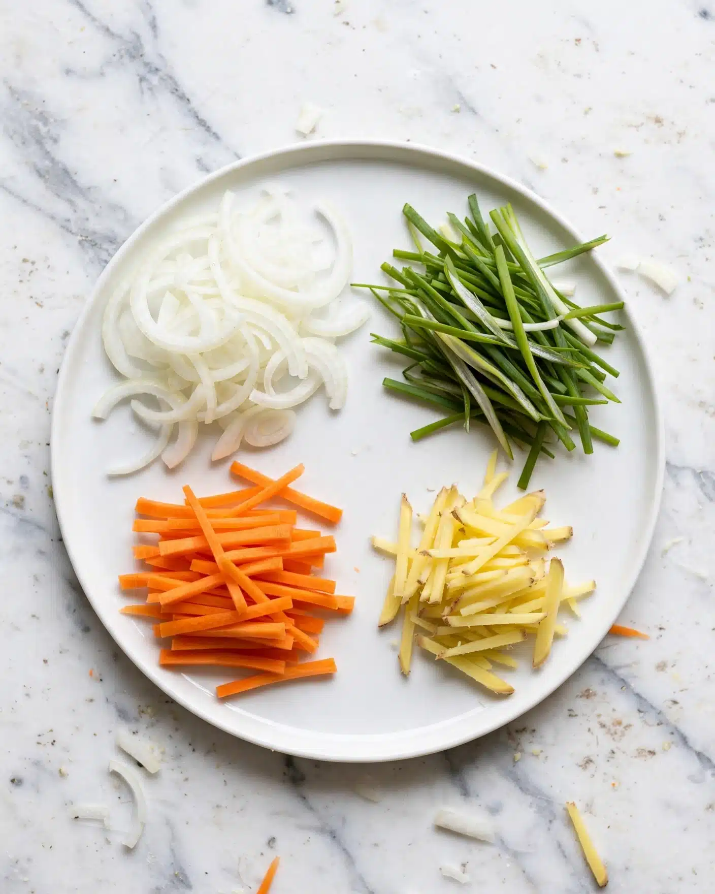 A white round plate sits on a white marbled surface, holding four separate piles of sliced vegetables. On the top left, there are thin white slices of onion with soft, curved edges. Below the onions, there is a pile of short, thin orange carrot sticks. On the bottom right, pale yellow thin sticks resembling ginger are neatly arranged. The top right section of the plate has a bunch of fresh green scallions, cut into long thin strips with some white parts visible. The colors are clear and fresh, giving a crisp and clean look. Photo taken with an iphone --ar 4:5 --v 7