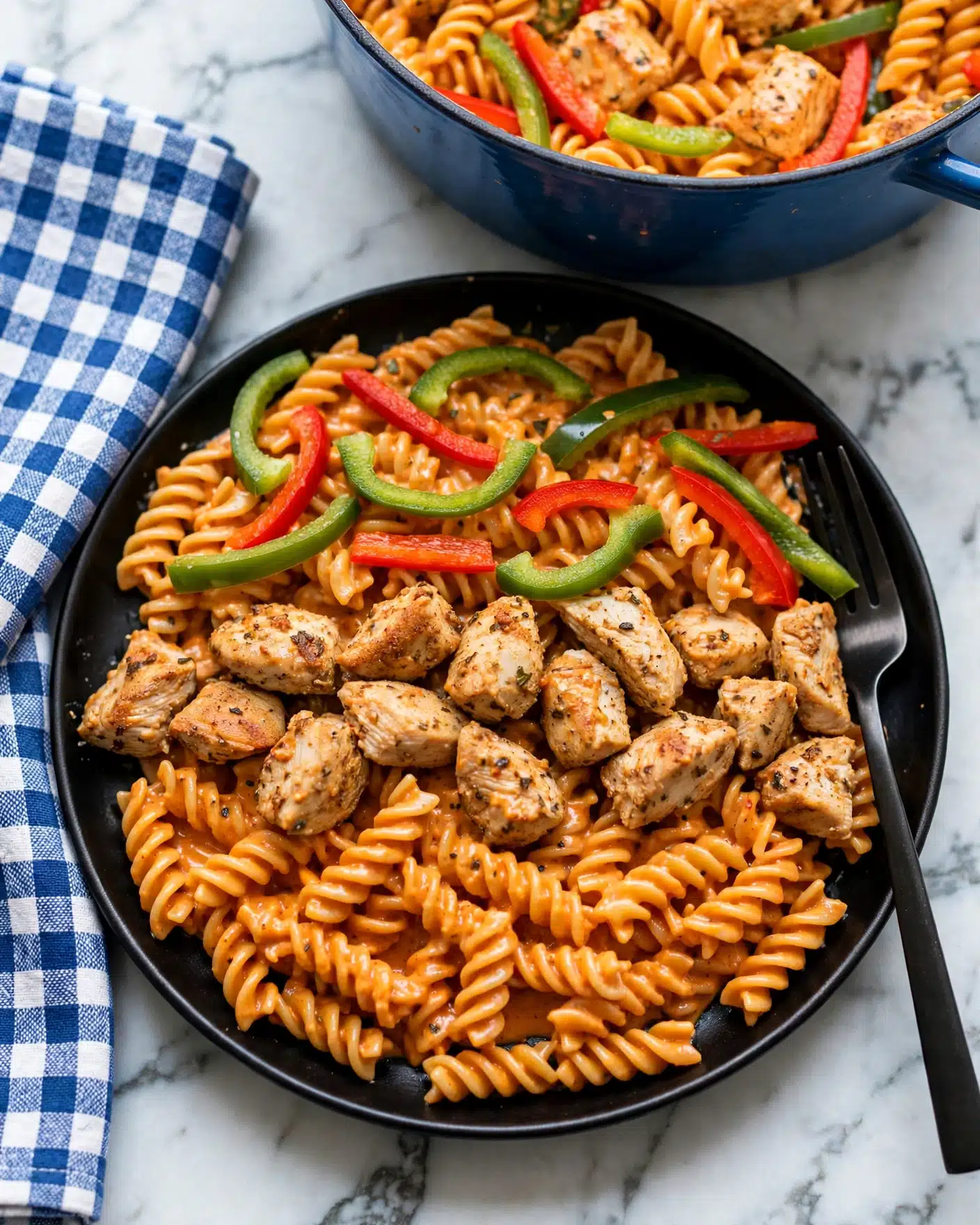 A close-up of a black plate filled with three layers: the base layer is orange spiral pasta coated in creamy sauce, the middle layer has chunks of light brown cooked chicken, and the top layer includes strips of green and red bell peppers scattered evenly. A dark fork rests on the right side of the plate. Above, a blue cooking pot filled with the same pasta mix is partially visible. The background is a white marbled surface with a folded blue and white cloth on the left side. Photo taken with an iphone --ar 4:5 --v 7