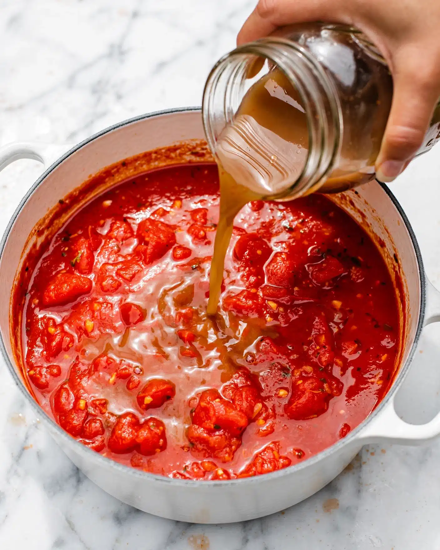 Roasted Eggplant and Tomato Soup Recipe 4 A close-up image shows a white pot filled with thick, bright red tomato sauce with some texture and small chunks visible on the surface. A woman's hand is pouring a light brown liquid from a clear mason jar into the sauce, creating a glossy area where the liquids meet. The pot's handles are visible on the clean white marbled surface. The lighting highlights the shine and richness of the sauce and liquid being poured. photo taken with an iphone --ar 4:5 --v 7