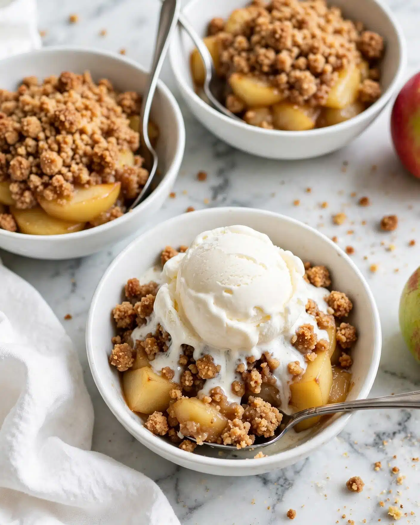 Healthy Apple Crumble Recipe 5 The image shows three white bowls placed on a white marbled surface. Each bowl contains a dessert with two visible layers: a bottom layer of soft, golden yellow cooked apple pieces mixed with a crumbly, brown streusel topping. On top of the dessert in the front bowl, there is a smooth, thick scoop of white vanilla ice cream, slightly melting and resting on the crumb layer. A silver spoon is placed in each bowl, with the spoon in the front bowl positioned under a portion of the crumble and apples. Some crumbs are scattered on the surface around the bowls. A woman’s hand is not visible interacting with the bowls, but a white cloth is present nearby along with a partially visible red and green apple. photo taken with an iphone --ar 4:5 --v 7