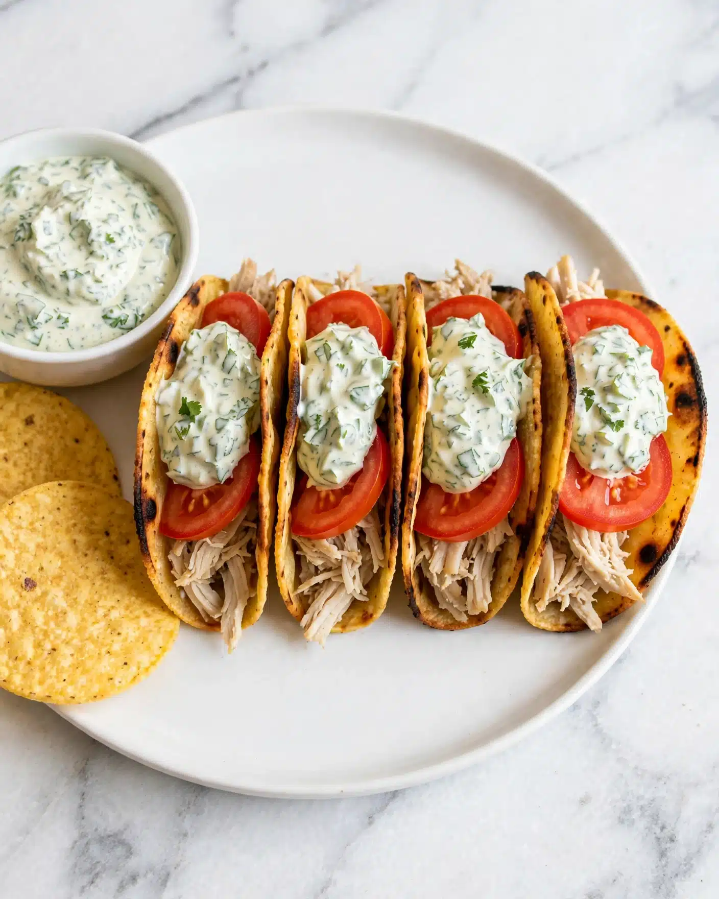 Four small tacos sit side by side on a white plate with a white marbled surface underneath. Each taco has a golden brown, lightly charred crispy shell filled with three main layers: the bottom layer is light beige shredded meat, next are bright red tomato slices, and the top layer is a creamy white sauce with green herbs mixed in. On the left side of the plate, there are two round yellow tortilla chips partially visible next to a small white bowl full of the same creamy herb sauce. Photo taken with an iphone --ar 4:5 --v 7