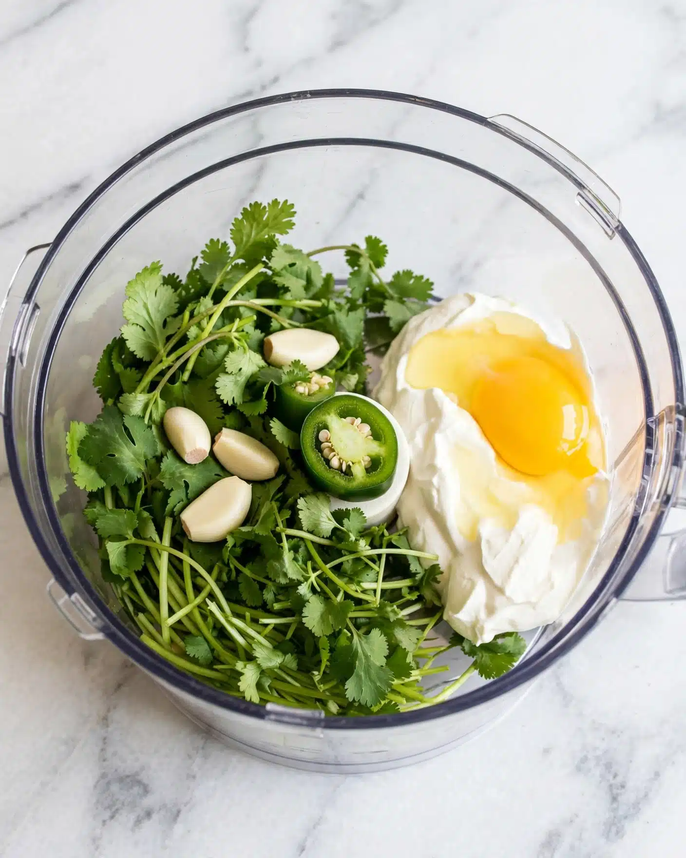 A clear round food processor bowl sits on a white marbled surface, filled with fresh green cilantro leaves and stems on one side, three pale garlic cloves scattered on top, and a halved green jalapeño pepper lying near the middle. On the other side, there is a dollop of white sour cream mixed with a light yellow egg yolk, all ready to be blended together. The layers show a mix of vibrant green herbs and vegetables with creamy white and pale yellow textures beside them, all contained within the transparent bowl. Photo taken with an iphone --ar 4:5 --v 7