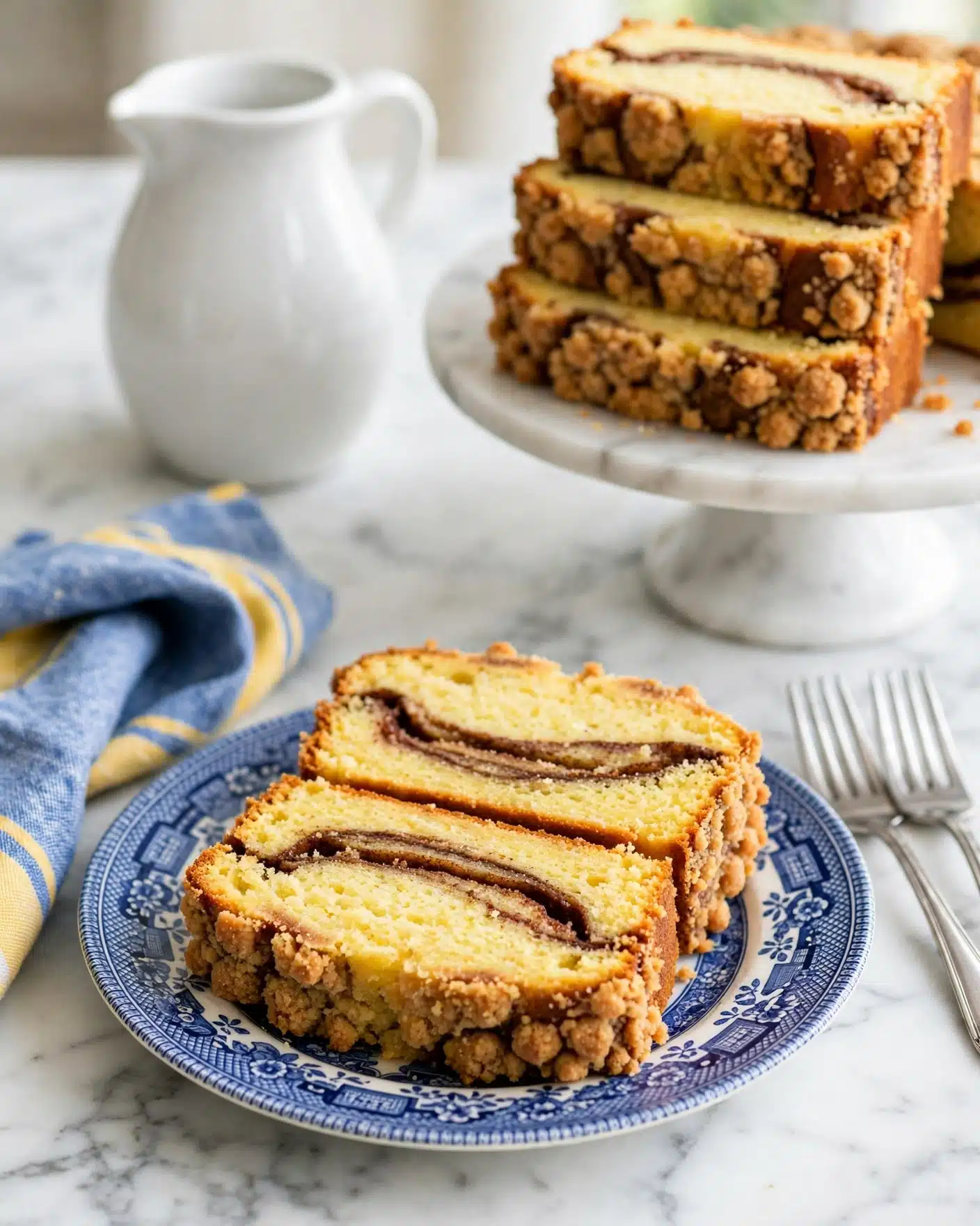 Decadent Cinnamon Swirl Bread Recipe 5 Two thick slices of yellow cake with a dark brown cinnamon swirl layer run through the middle, placed on a blue and white patterned plate. The cake has a crumbly golden-brown crust on the sides. Behind the plate, there is a white ceramic jug and a white marble cake stand holding more slices of the same cake. The surface is a white marbled texture, with a folded blue and yellow napkin to the left and two silver forks to the right. Photo taken with an iphone --ar 4:5 --v 7