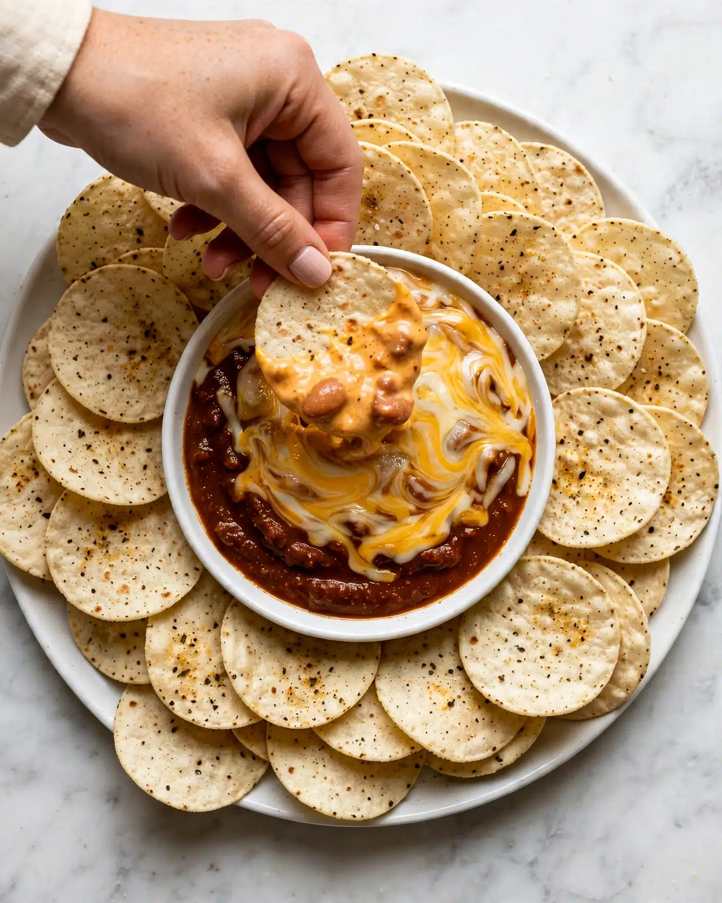 A woman's hand holds a round, light beige tortilla chip dipped halfway into a bowl of thick, creamy orange cheese dip with visible bits of melted cheese and spices. The white bowl containing the dip is placed on a white plate, which is fully covered with many tortilla chips arranged in a circular, overlapping pattern. The background shows a white marbled texture. photo taken with an iphone --ar 4:5 --v 7