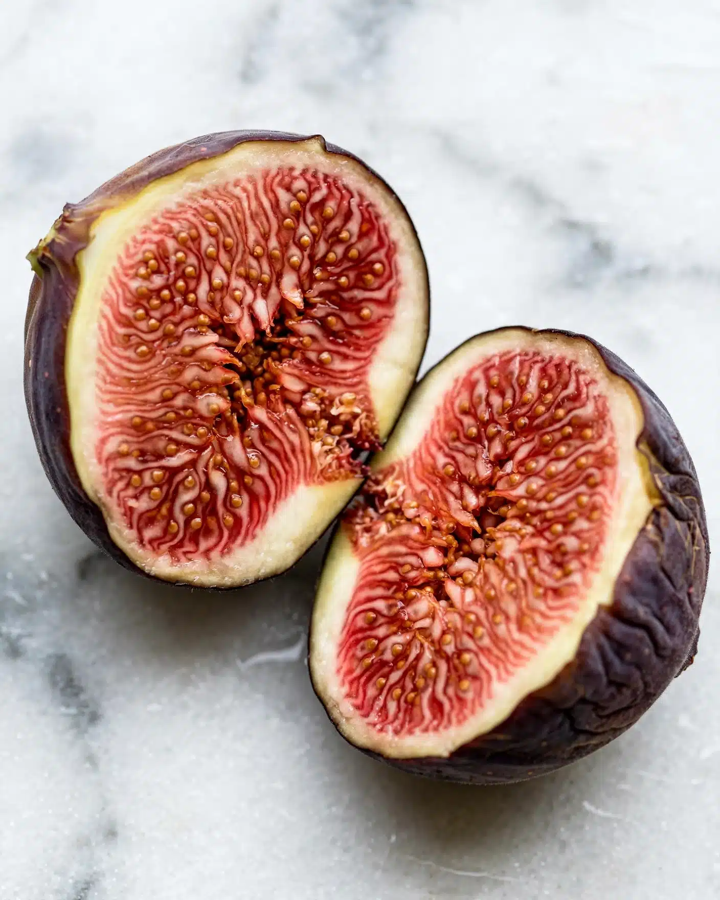 The image shows two halves of a ripe fig placed on a white marbled surface. Each fig half reveals a soft, textured inside with a pinkish-red color that has tiny seeds and a slightly fibrous look. The outer skin of the fig is a deep purple-brown color, smooth but slightly wrinkled, providing a contrast to the vibrant inner part. The figs are positioned close to each other, highlighting the juicy, fresh appearance of the fruit inside. photo taken with an iphone --ar 4:5 --v 7