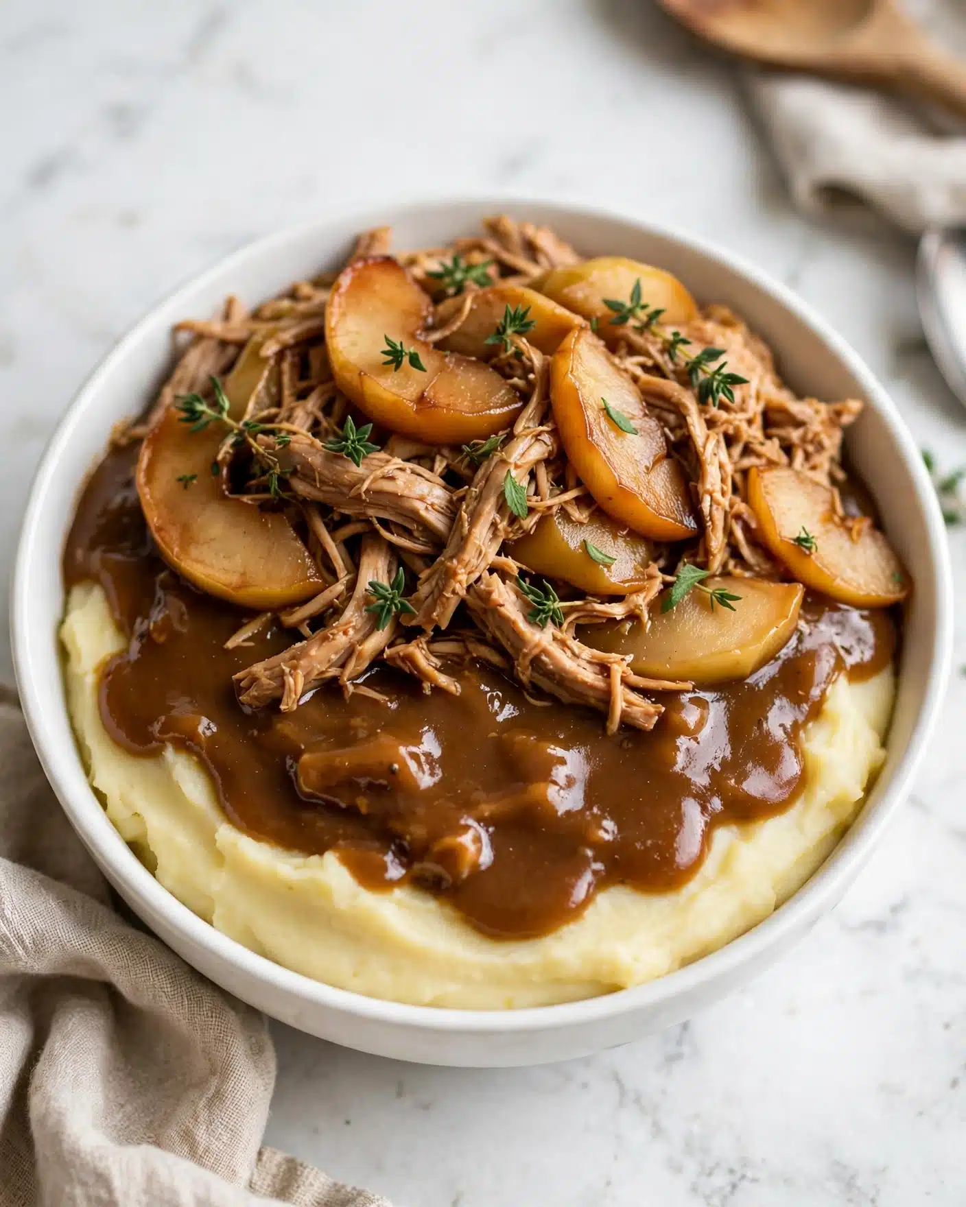 The image shows a round white bowl filled with three layers. The bottom layer is smooth pale yellow mashed potatoes, spread evenly to cover the base. On top of this is a thick brown gravy with a glossy texture, partially covering the mashed potatoes. The top layer consists of tender, shredded chicken mixed with cooked, sliced apples and small sprigs of fresh green thyme scattered over the dish. The bowl is set on a white marbled surface with a beige kitchen towel and some kitchen items slightly visible around it. Photo taken with an iphone --ar 4:5 --v 7
