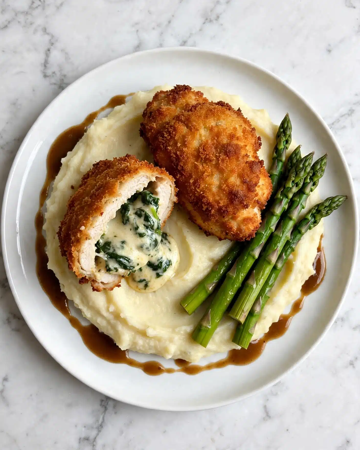 Crispy Homemade Chicken Kiev with Herb Butter Recipe 5 A white plate sits on a white marbled surface holding creamy, smooth mashed potatoes forming the base layer, topped with golden-brown crispy breaded pieces filled with green spinach and white cheese at the center, one piece is whole and the other is cut in half revealing the filling. On the side, there are bright green cooked asparagus spears resting against the mashed potatoes. A glossy brown gravy is gently drizzled around the edges of the mashed potatoes. Photo taken with an iphone --ar 4:5 --v 7