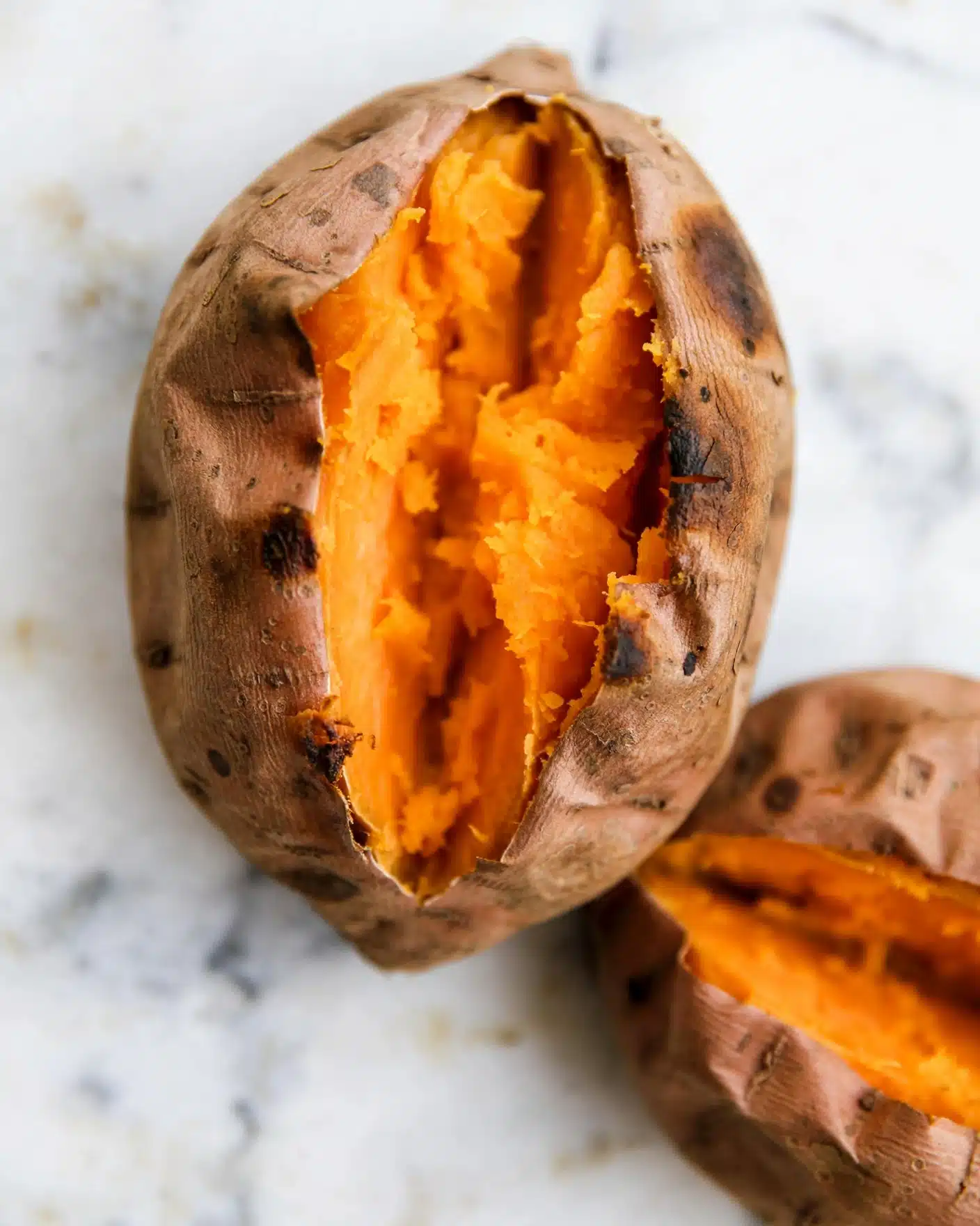 Two baked sweet potatoes sit on a surface with white marbled texture. Each sweet potato is whole but has a wide split down the middle, revealing a soft, bright orange interior that looks tender and cooked through. The skin is brownish with some wrinkles and small dark spots, showing natural texture and slight caramelization. The close-up view focuses mostly on the top sweet potato, while the second one is partially visible in the background. Photo taken with an iphone --ar 4:5 --v 7