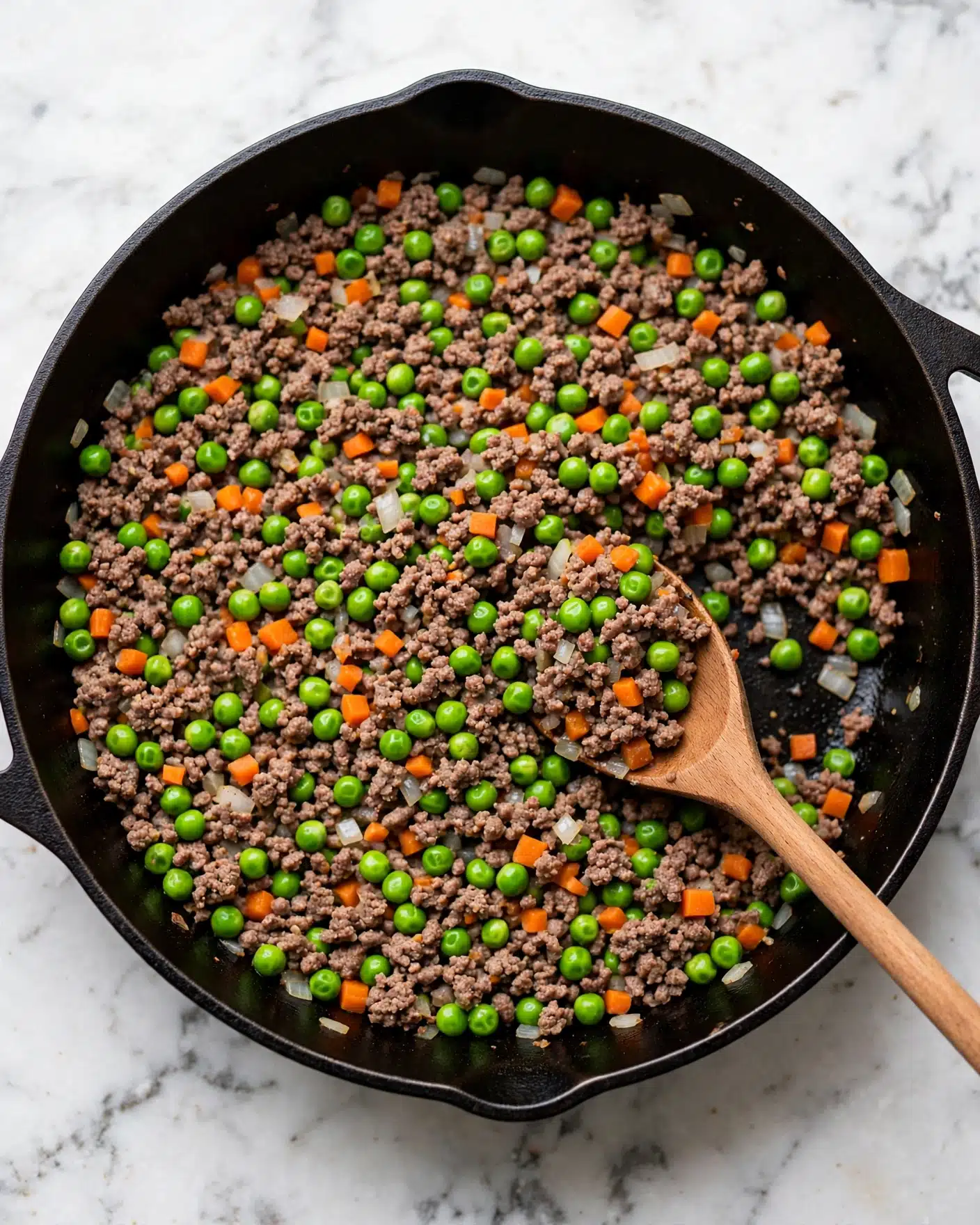 The image shows a close-up of cooked ground beef mixed with bright green peas, small orange carrot pieces, and small white onion bits, all evenly spread in a black pan. A light brown wooden spoon is placed inside the pan, slightly lifting some of the mixture, with the spoon handle visible and pointing towards the bottom right. The texture of the cooked ingredients looks soft but distinct, with a mix of small round peas and tiny carrot cubes adding color contrast. The background is a white marbled texture. Photo taken with an iphone --ar 4:5 --v 7