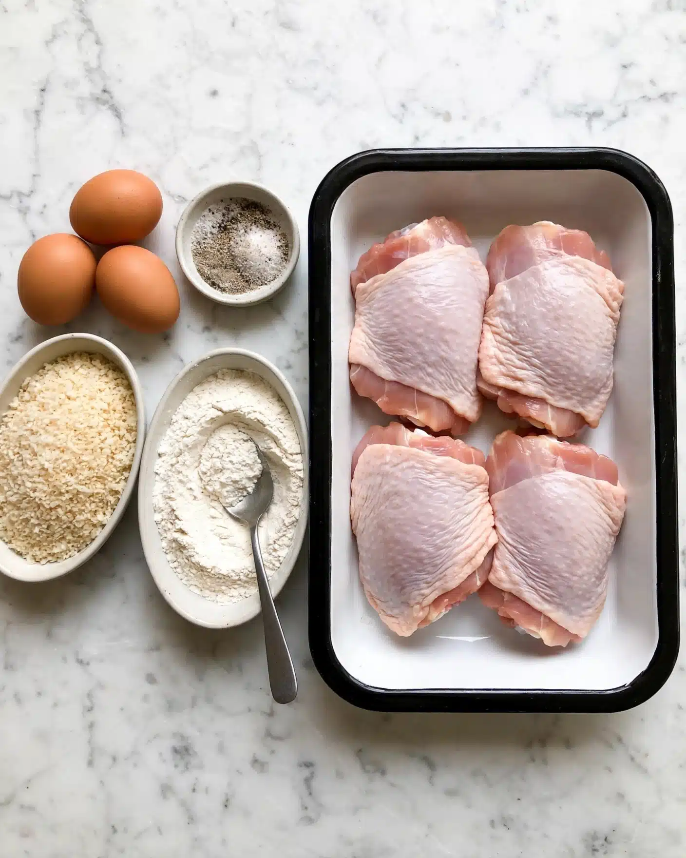 Crispy Homemade Chicken Kiev with Herb Butter Recipe 4 The image shows a white tray with a black rim holding four raw pink chicken thighs arranged close together. To the left of the tray, three brown eggs rest on a white marbled surface, above a small round bowl filled with a mixture of salt and pepper. Below the eggs, there is an oval bowl filled with light beige panko crumbs, and to its right, a larger oval bowl contains white flour with a spoon resting inside. The whole setup is laid out on a white marbled surface. Photo taken with an iphone --ar 4:5 --v 7