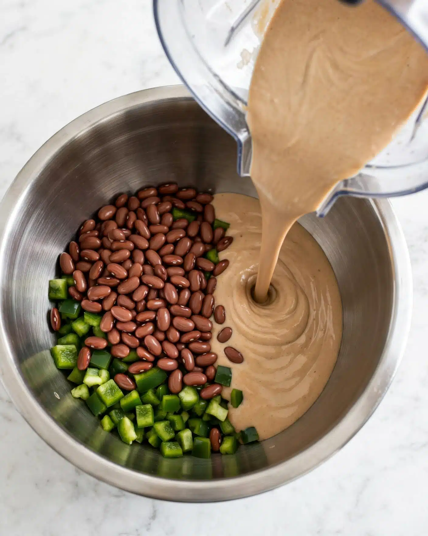 A stainless steel mixing bowl sits on a white marbled surface, filled partially with a layer of whole brown beans on one side and chopped green peppers visible beneath. A thick, creamy light brown sauce is being poured from a clear blender container into the bowl over the other half of the ingredients, showing a contrast between the smooth sauce and the firm beans. The bowl's shiny metal interior reflects some soft light, and the background is clean and bright. photo taken with an iphone --ar 4:5 --v 7